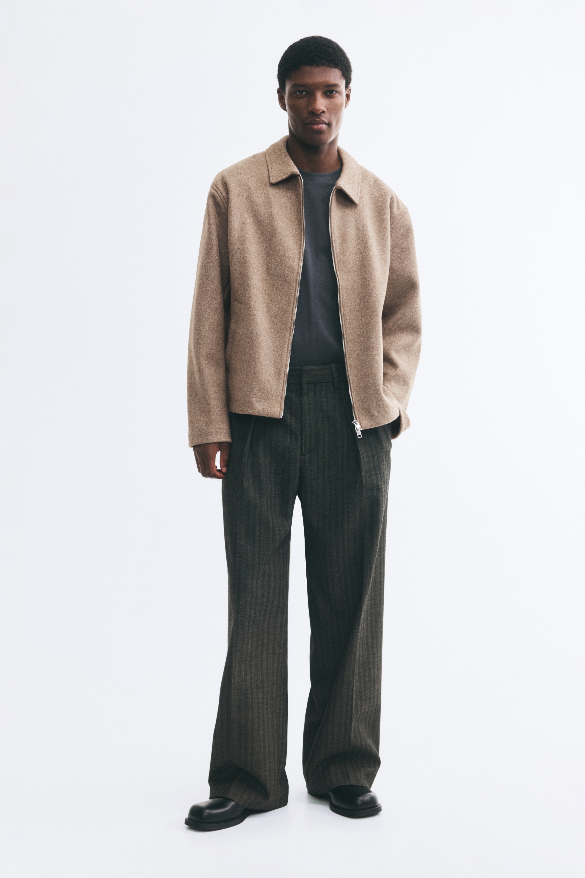 La veste marron clair est présentée de face, mettant en valeur un tissu texturé. Elle arbore une fermeture éclair argentée sur toute la longueur, un col classique à pointes écartées et deux poches passepoilées en biais sur le bas du devant.