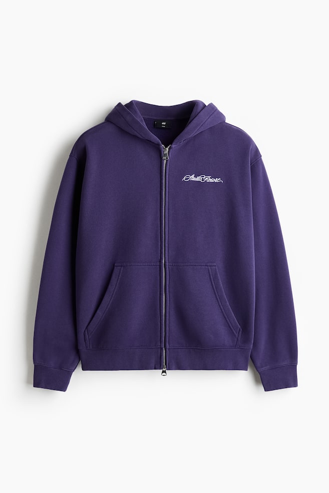 Ein tiefvioletter Kapuzenpullover mit Reißverschluss ist von vorne zu sehen und zeigt einen silberfarbenen Reißverschluss in voller Länge sowie eine angenähte Kapuze. Er verfügt über eine Kängurutasche und den weißen, gestickten Schriftzug "Studio Resort" auf der linken Brust, mit gerippten Bündchen und einem gerippten Saum. - 1