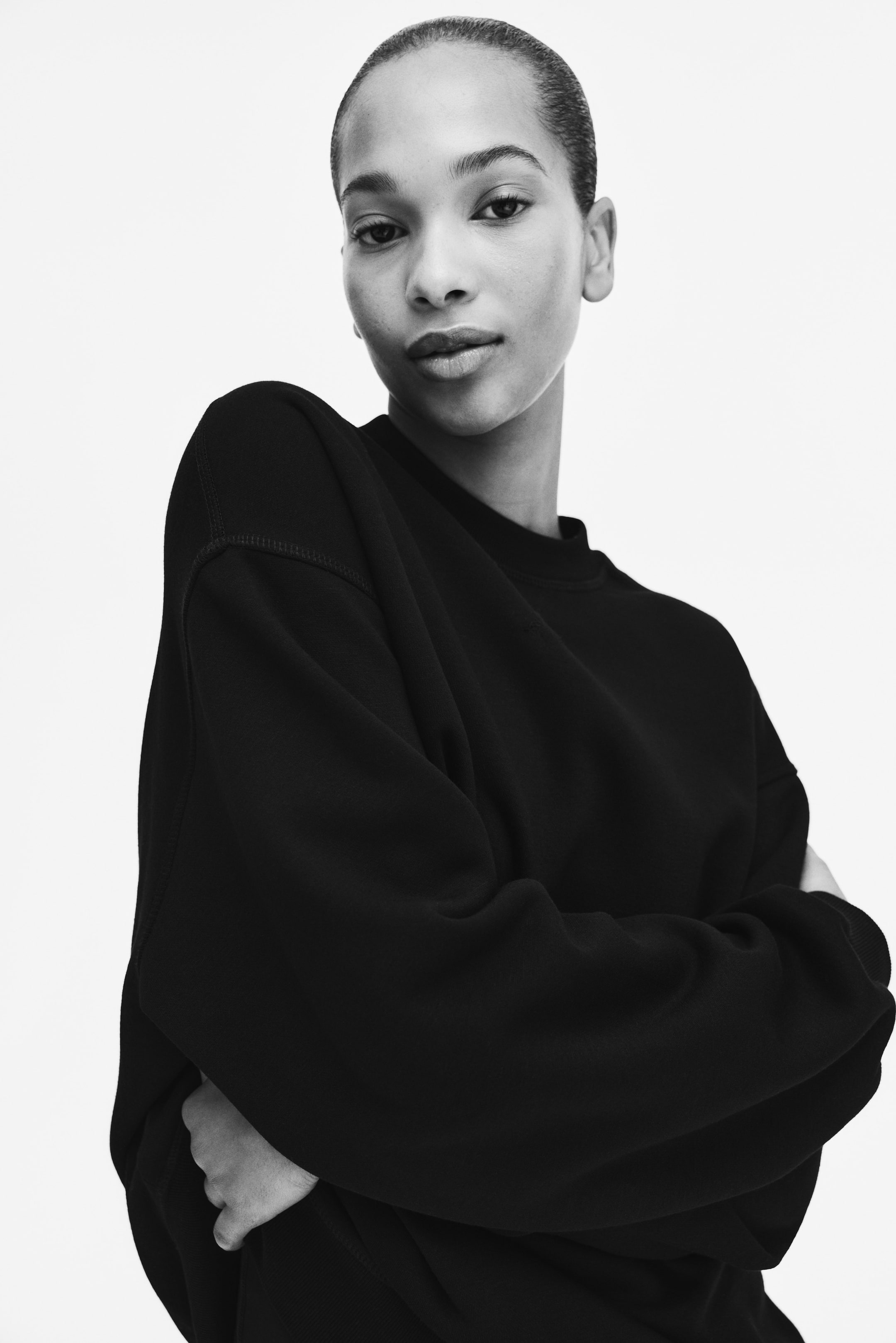 Agrandir l'image: Un sweat-shirt noir à col rond présente une coupe décontractée avec des manches raglan et des coutures apparentes le long de la couture de l'épaule. Les manches semblent amples alors que les bras du mannequin sont croisés devant.