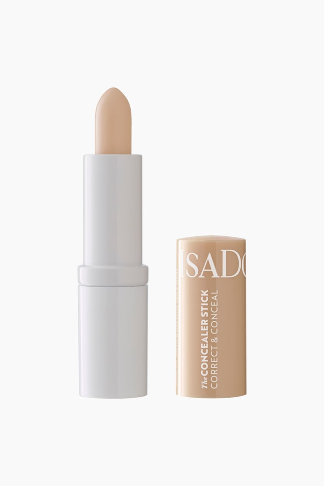 En lys beige concealerstift står oppreist, produktet er delvis ført ut av den hvite, sylindriske basen og danner en spiss tupp. Den matchende lyse beige hetten med hvit tekst ligger ved siden av. - 1