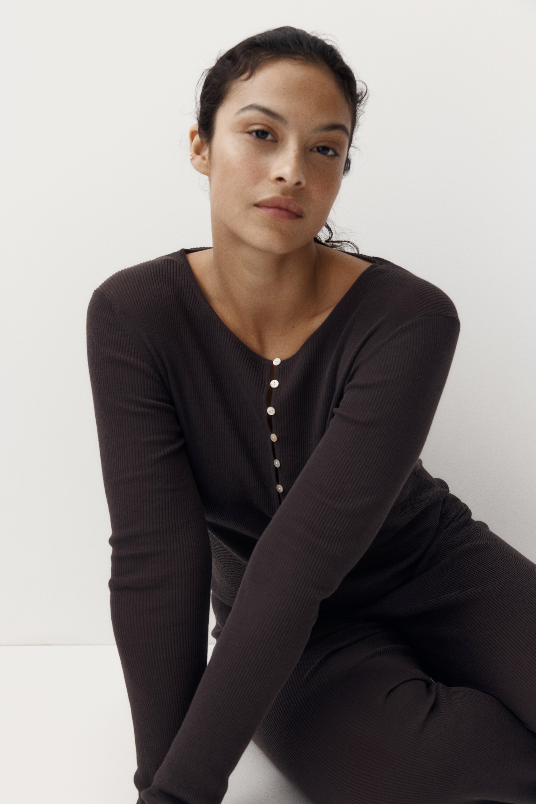 View larger image: "A dark brown ribbed knit top paired with matching pants creates a sleek silhouette. The top features a henley neckline with a partial button placket, adorned with small light-coloured buttons, and long sleeves."