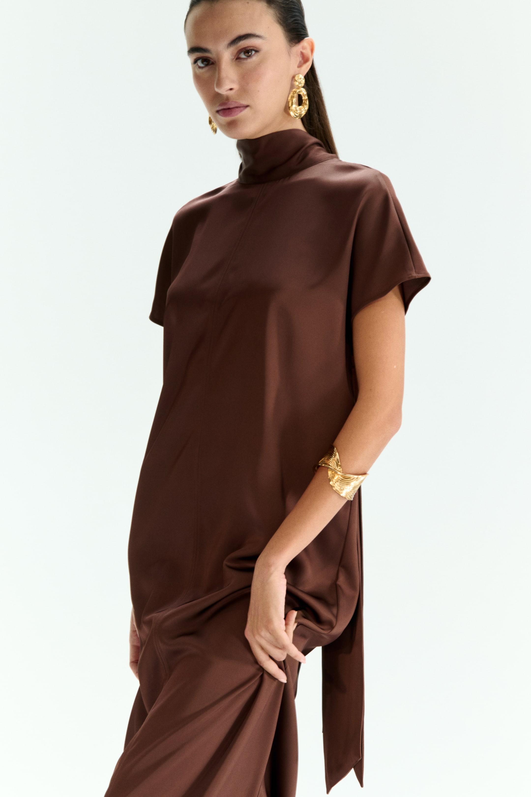Agrandir l'image: Une robe marron à col montant avec des manches courtes est portée par le mannequin, qui rassemble doucement le tissu d'une main. Des boucles d'oreilles en or et un bracelet manchette en or accessoirisent la robe.