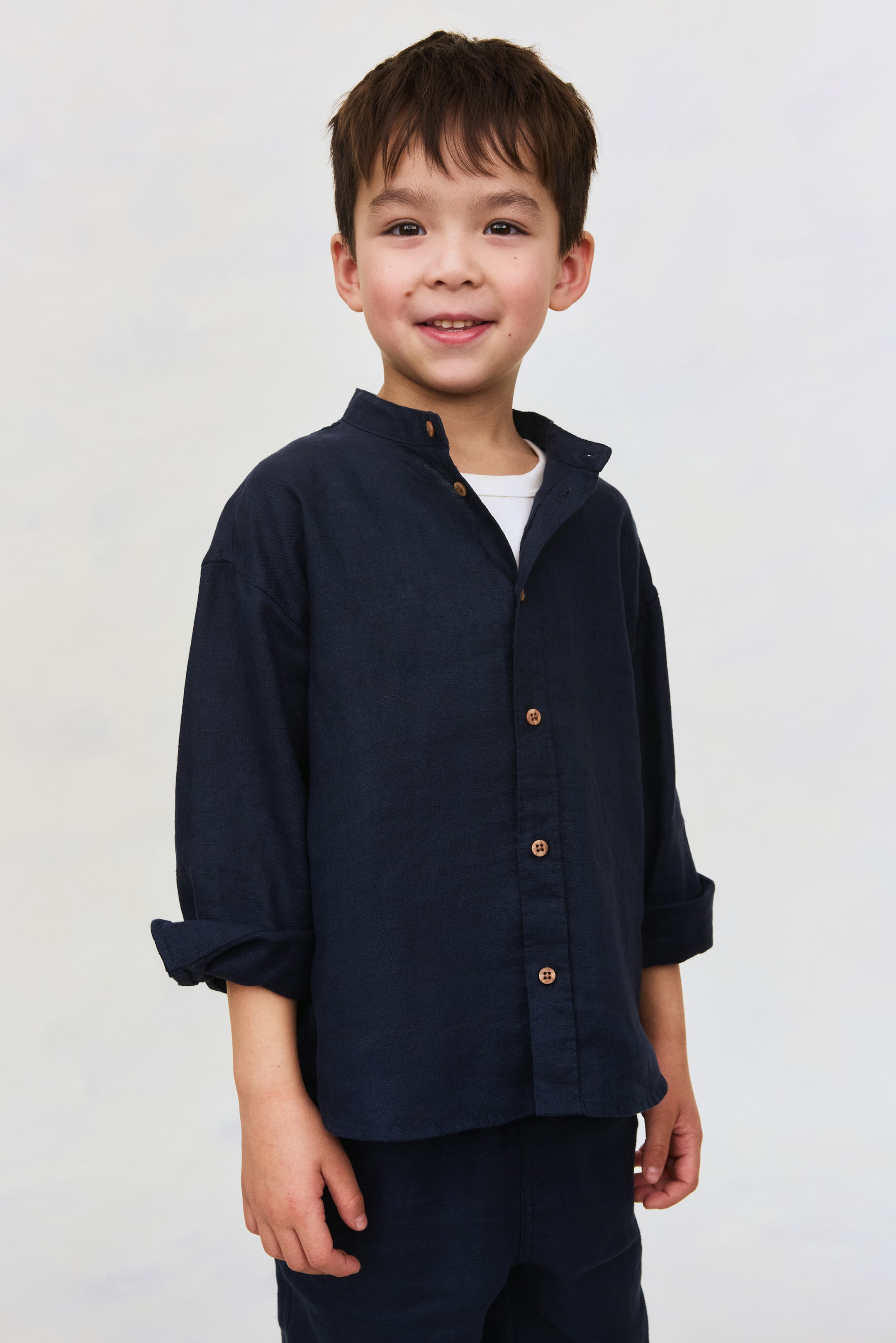View larger image: A dark blue linen shirt is shown from the front, featuring a band collar, light brown buttons running down the front, and dropped shoulders with long sleeves rolled up at the cuffs; a white T-shirt is visible beneath the open neckline.