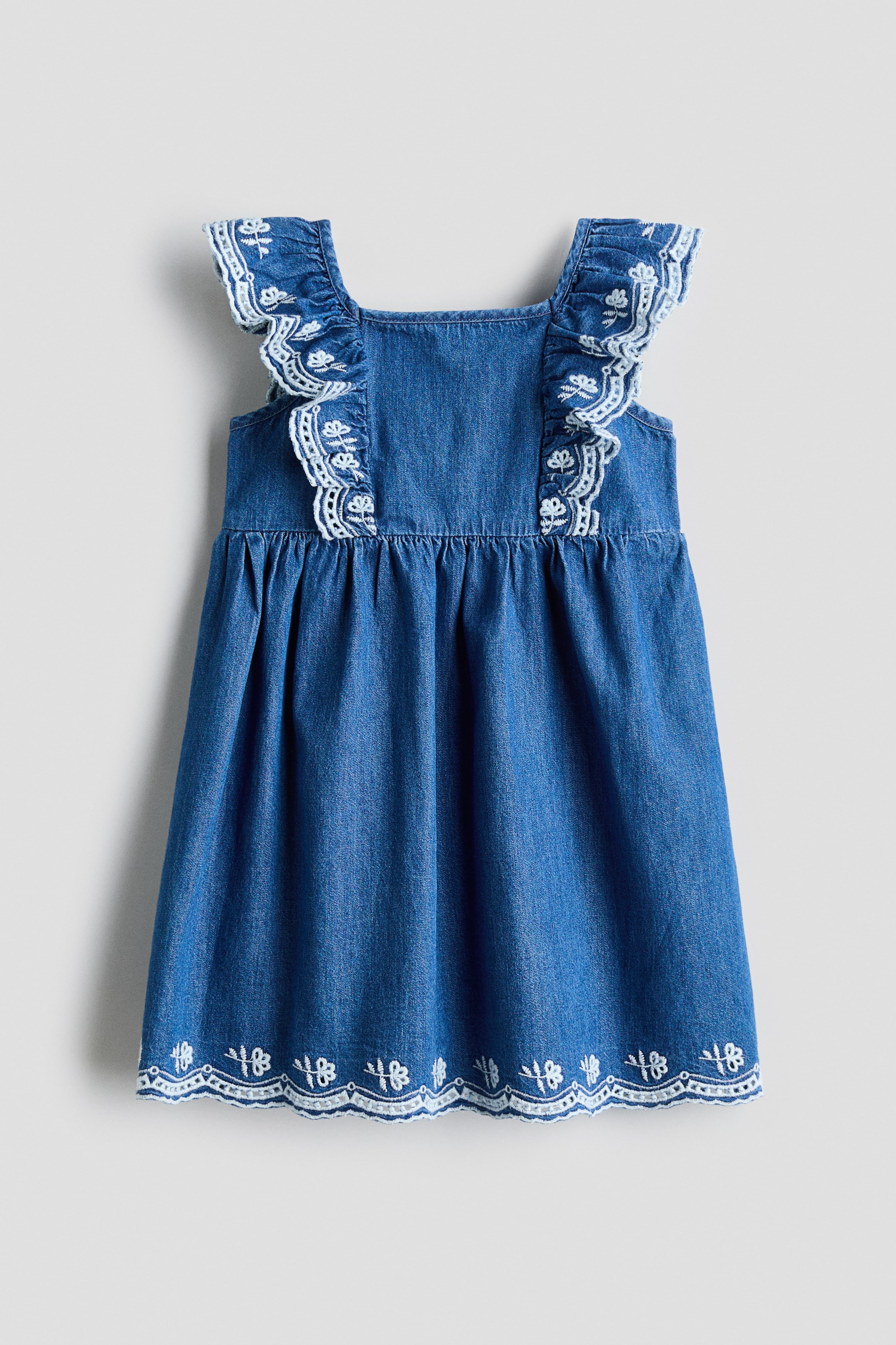 View larger image: The flared, sleeveless cotton denim dress is showcased from the front. It features a square neckline, with flounces adorned with white embroidered motifs extending over the shoulders. A gathered seam defines the waist, and the scalloped hem is detailed with a matching embroidered border.