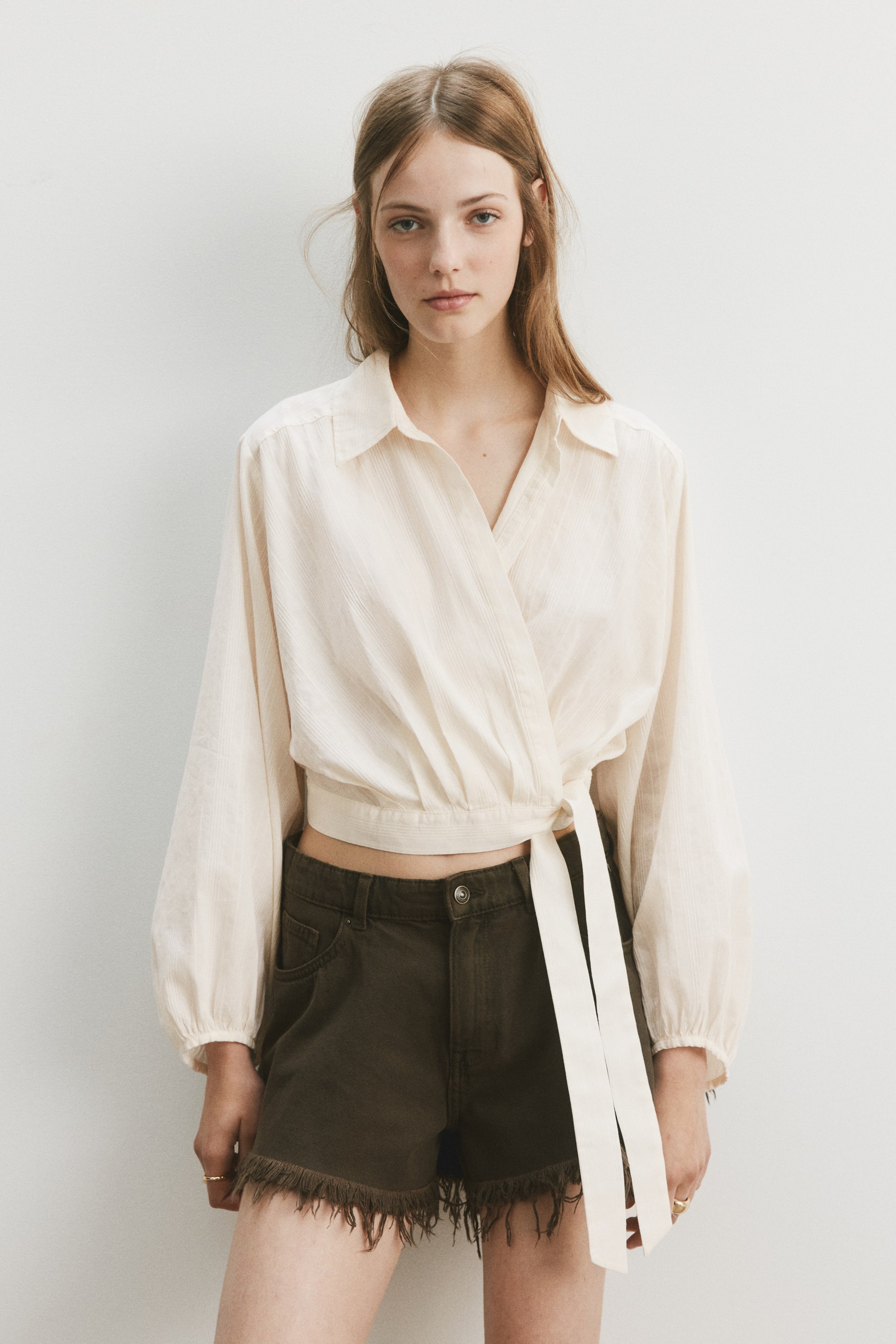 View larger image: The off-white wrap top showcases a subtle vertical stripe texture, featuring a pointed collar and a V-neckline. Its long, voluminous sleeves gather into shirred cuffs, and the wrap design ties at the side waist, creating a cropped silhouette.