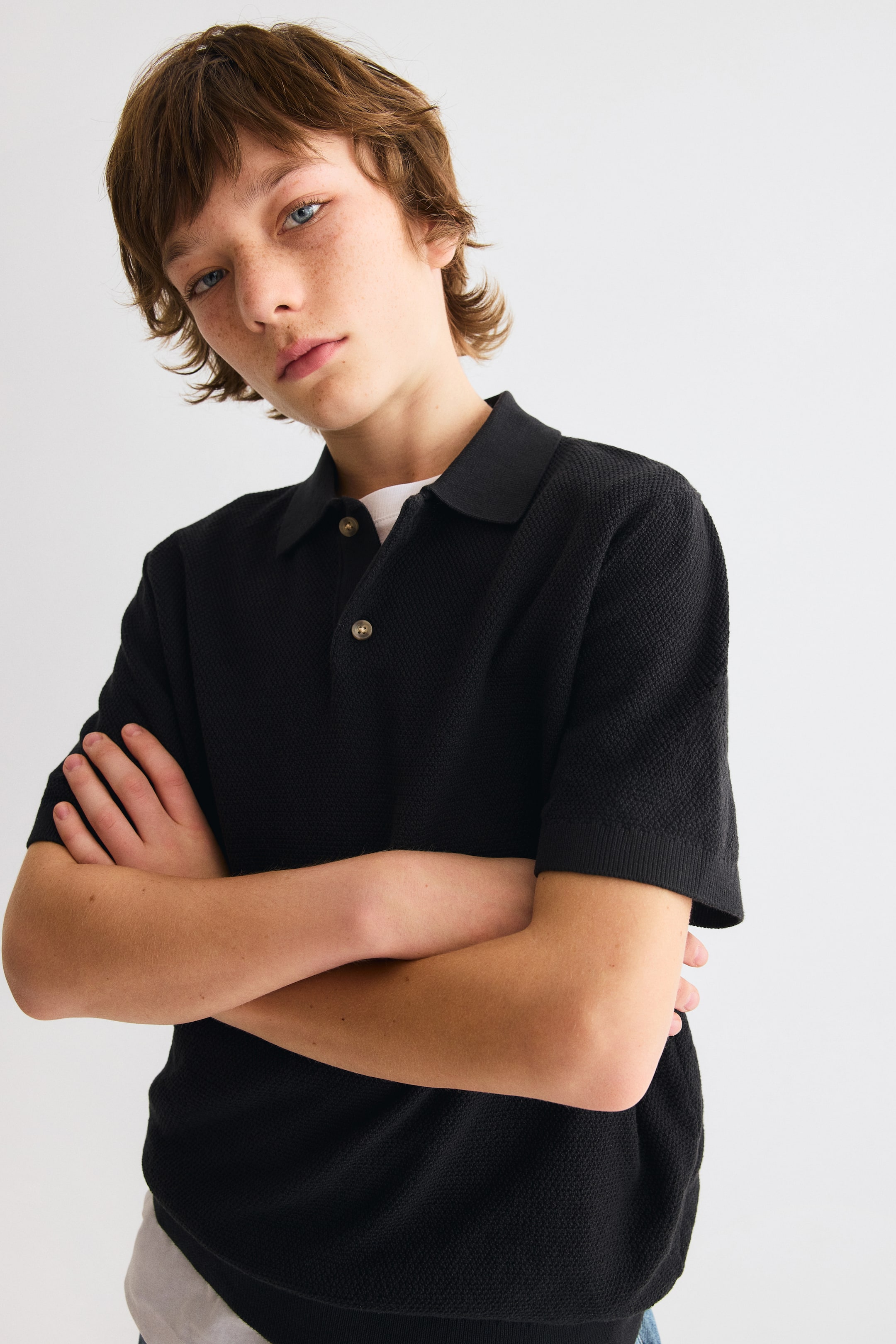 Ver imagem maior: Uma camisa polo preta está a ser apresentada de um ângulo ligeiramente frontal-esquerdo, com um tecido de malha texturizada, uma gola clássica e uma carcela com dois botões castanho-claros. Apresenta punhos canelados nas mangas curtas e uma bainha canelada, com uma camisola interior branca visível na gola.