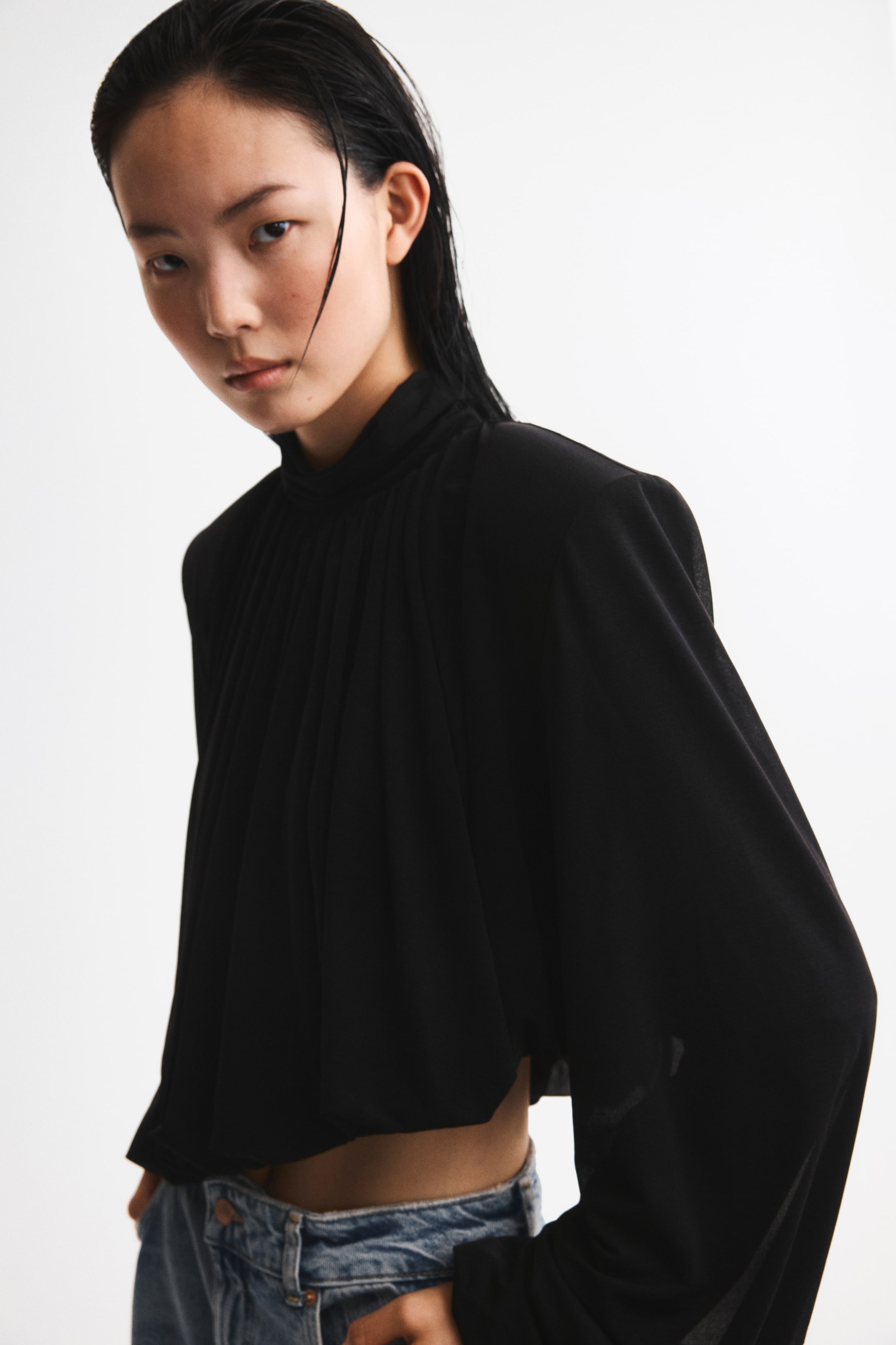 View larger image: The model is wearing a black cropped top. It features a gathered turtleneck, shoulder pads, and long balloon sleeves with elasticated cuffs. The top has an elasticated bubble hem, creating a puffy silhouette, and is styled with blue jeans.