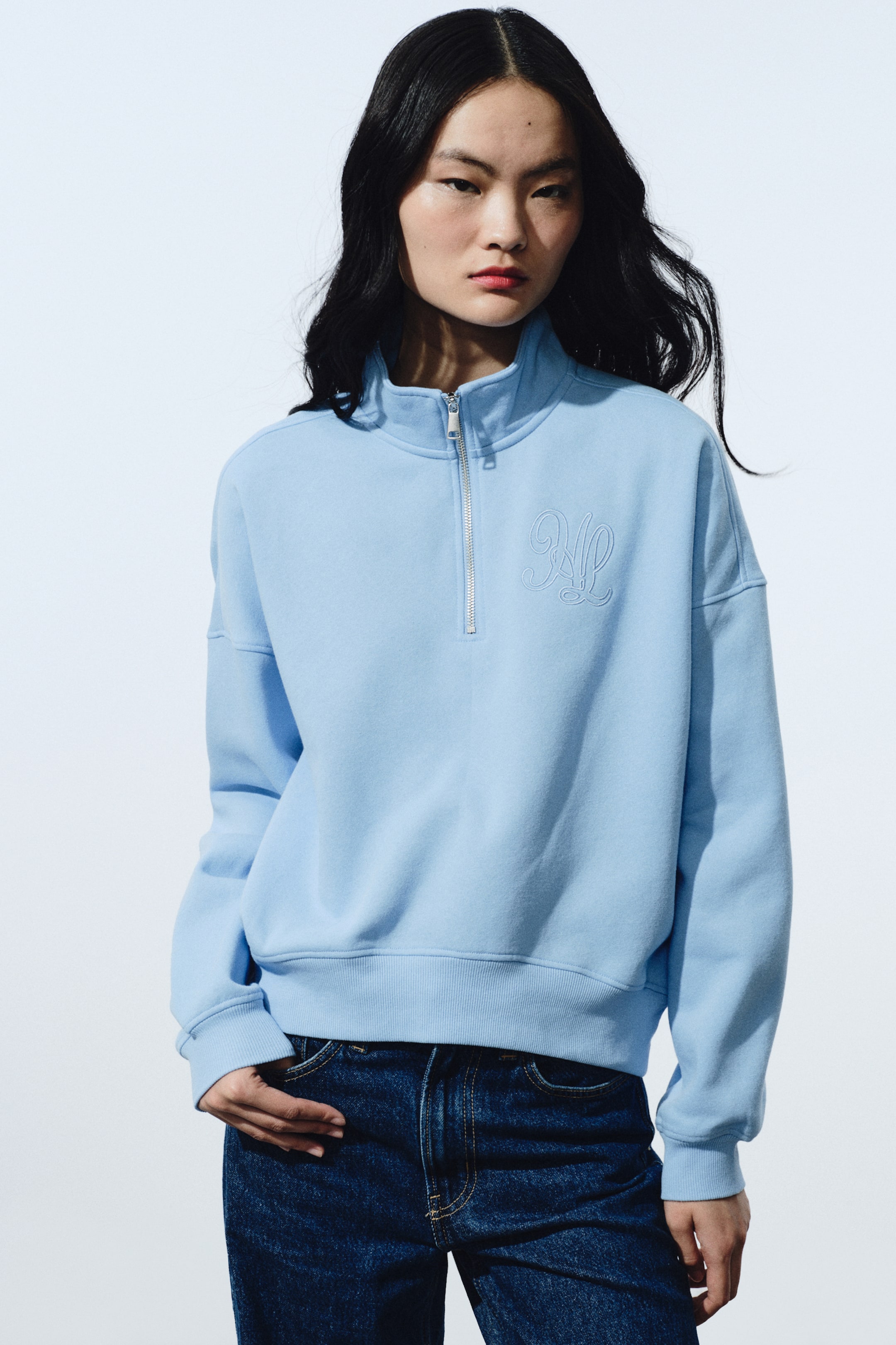 拡大画像を表示: ライトブルーのクォータージップスウェットは、スタンドカラー、シルバートーンのクォータージップ、左胸の刺繍モノグラムが特徴です。リブ仕上げの袖口と裾を備え、ダークブルーのジーンズの上でヒップにかかる丈になっています。