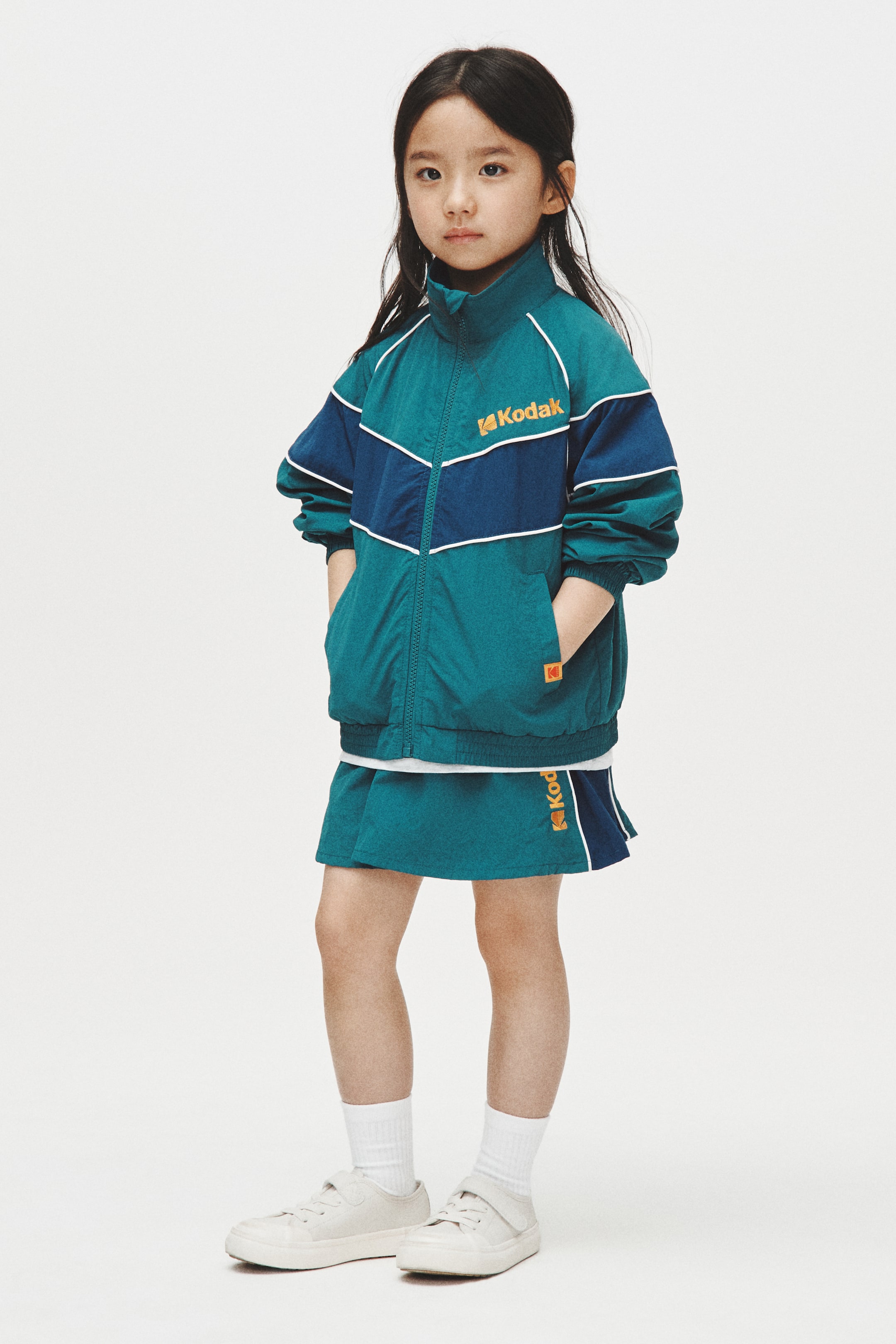 이미지 확대 보기: 청록색과 네이비 블루로 구성된 컬러 블록 트랙수트가 오른쪽을 약간 향한 재킷과 매칭되는 스커트를 선보이고 있습니다. 재킷은 전체 지퍼 클로저, 스탠드 칼라, 리브드 커프스와 밑단, 청록색과 네이비 부분을 강조하는 흰색 파이핑, 가슴에 오렌지색 코닥 로고와 앞주머니에 K 로고를 특징으로 하고 있습니다. 매칭되는 스커트는 네이비 블루 패널, 흰색 파이핑, 오렌지색 코닥 로고를 보여주고 있습니다.