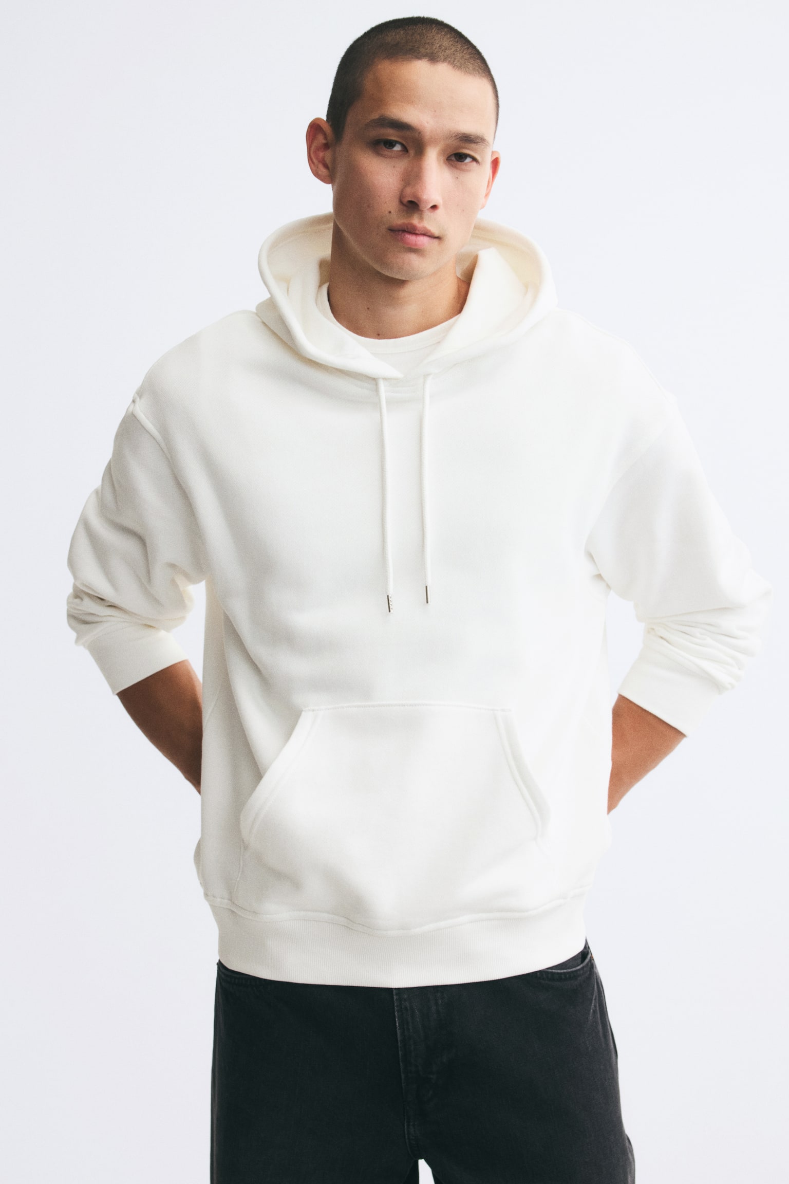 The model is wearing an off-white hoodie featuring a relaxed fit, drawstring hood, dropped shoulders, a kangaroo pocket, and a ribbed hem. The long-sleeved hoodie is styled with black jeans. - 1