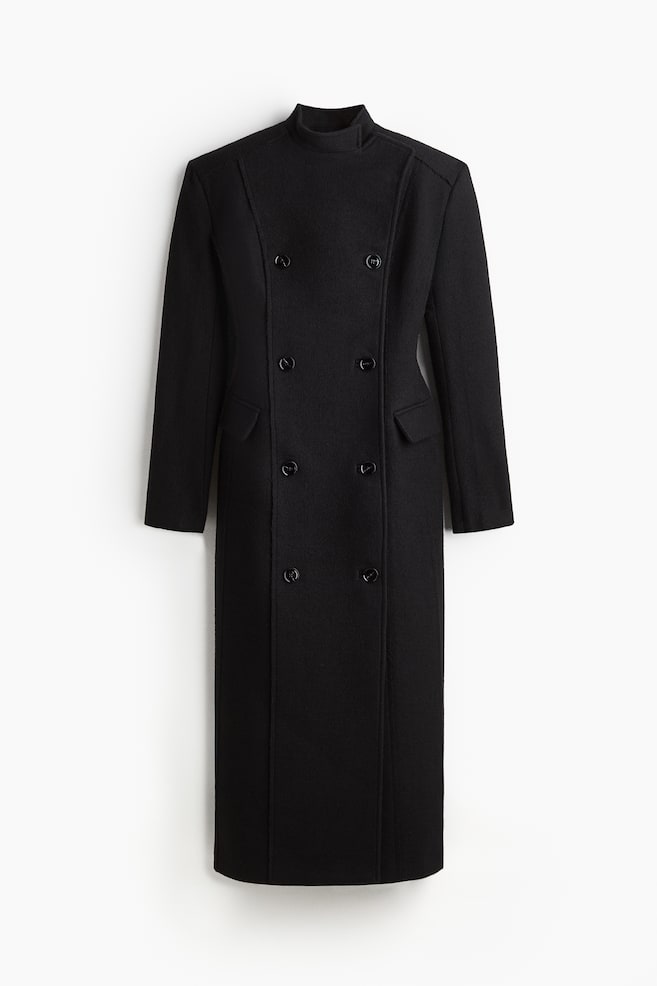 A black, ankle-length double-breasted coat is being presented from the FRONT, showcasing a structured fit and sharp silhouette. It is featuring a stand collar, prominent vertical seams, and six visible buttons arranged in two vertical rows. Jetted front pockets with flaps are also visible, along with long sleeves and a straight-cut hem. - 1