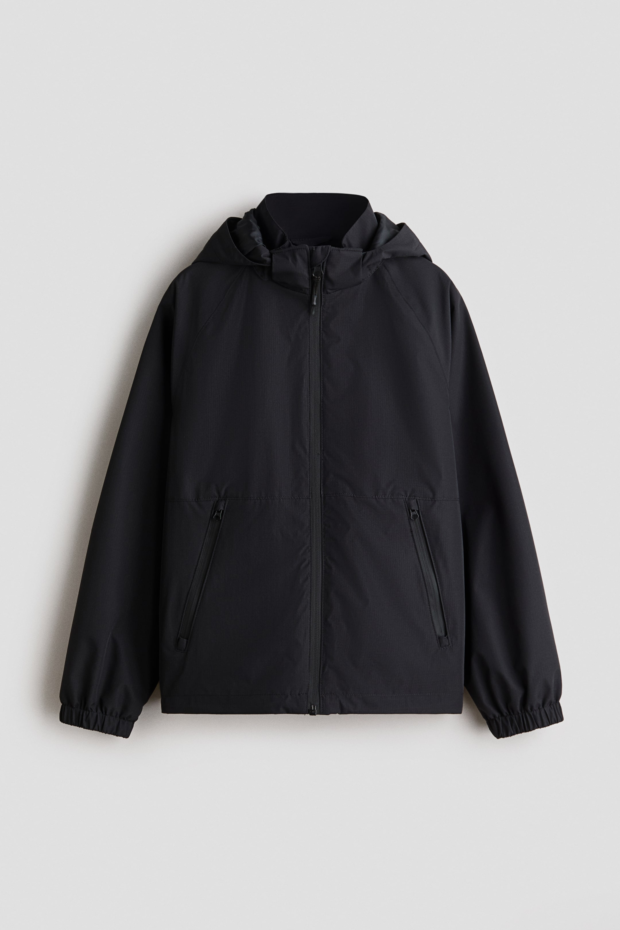 View larger image: A black shell jacket is presented front-facing, featuring a detachable hood, a stand-up collar, and a full zip closure. It is designed with raglan sleeves, two zipped side pockets, elasticated cuffs, and a drawstring-adjustable hem.
