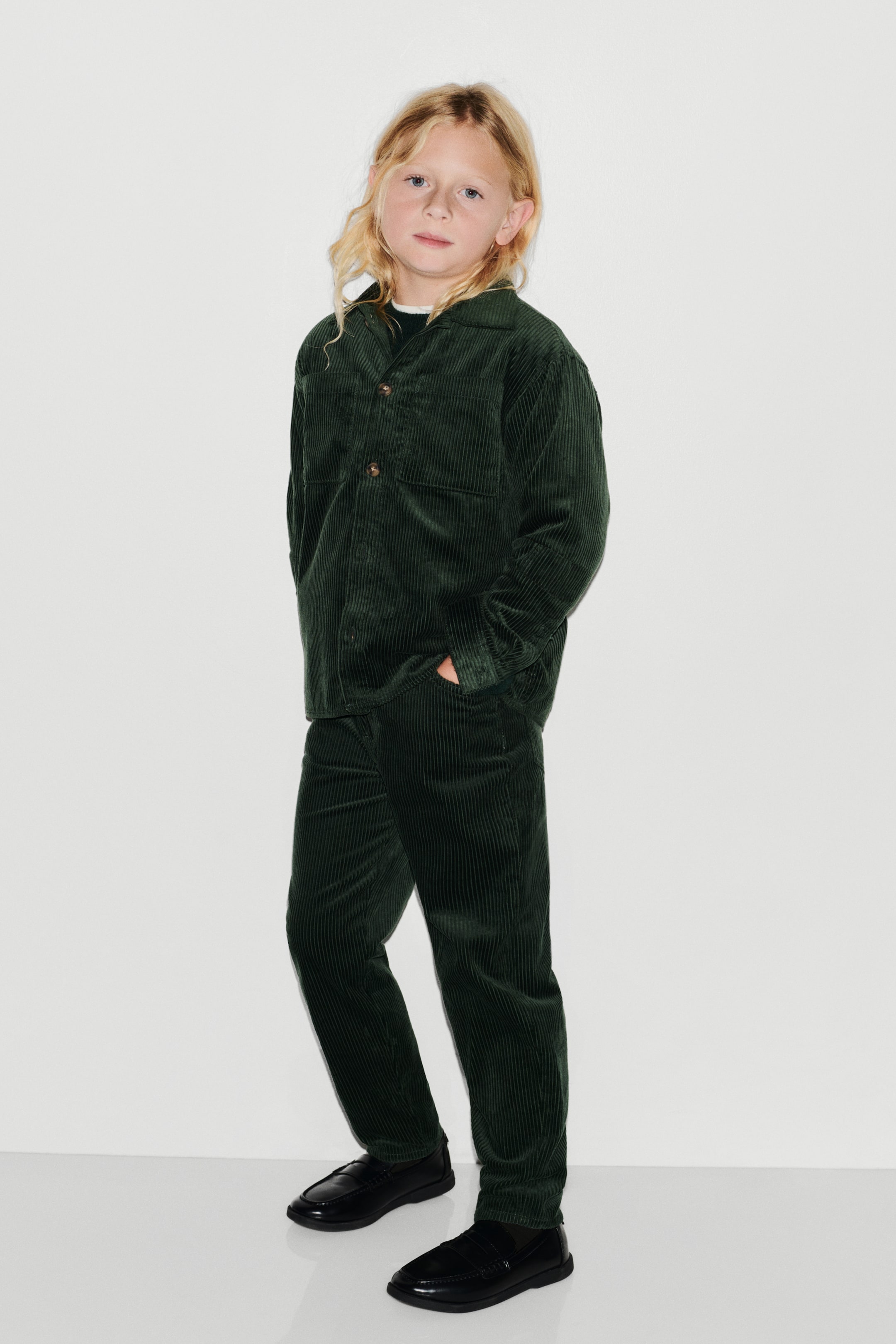 Agrandir l'image: Ce pantalon en velours côtelé vert foncé présente une coupe ample au niveau des hanches, des cuisses et des mollets, avec un tombé légèrement orienté vers la droite pour mettre en valeur sa silhouette décontractée à jambe droite. Il est associé à une surchemise assortie en velours côtelé vert foncé et à des mocassins noirs, soulignant son style casual.