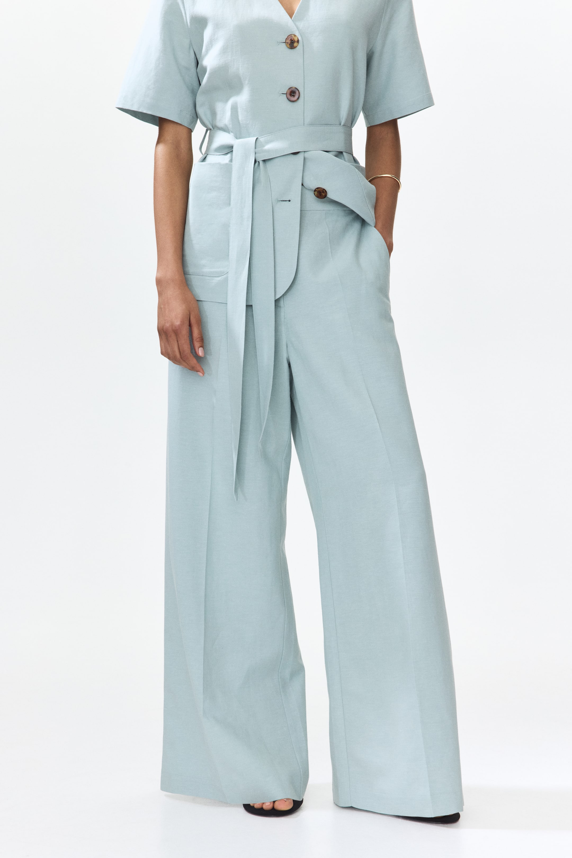 View larger image: "Light blue, loose-fit trousers are showcased from the front, highlighting a high waist with a coordinating fabric belt tied around the waist. These trousers feature diagonal front pockets and wide legs with prominent creases running down the front."