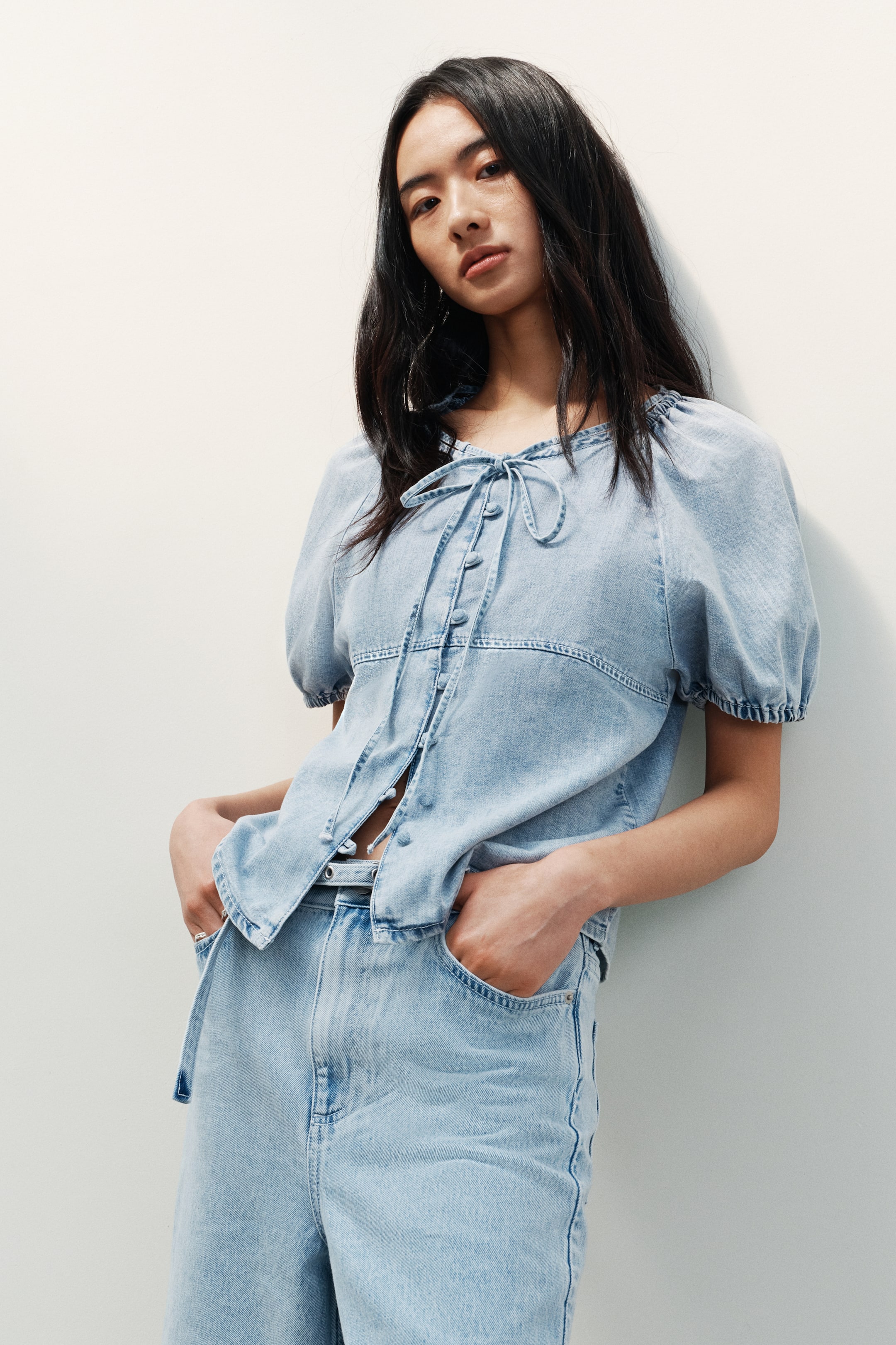 View larger image: A light blue denim top dan seluar lebar yang sepadan sedang dipamerkan. Top ini menampilkan leher berkerut dengan penutup tali, lengan pendek yang kembung, dan hadapan berkancing dengan beberapa butang bawah tidak diikat. Seluar berpinggang tinggi ini menampilkan poket sisi dan potongan yang santai.