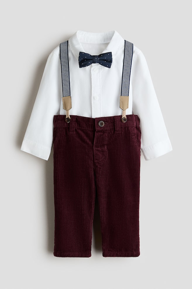 A childrenâs clothing set is showing a front view of a white long-sleeved button-up shirt, styled with a navy-blue polka-dot bow tie. The shirt is layered under striped suspenders that are buttoned onto deep burgundy corduroy trousers. - 1