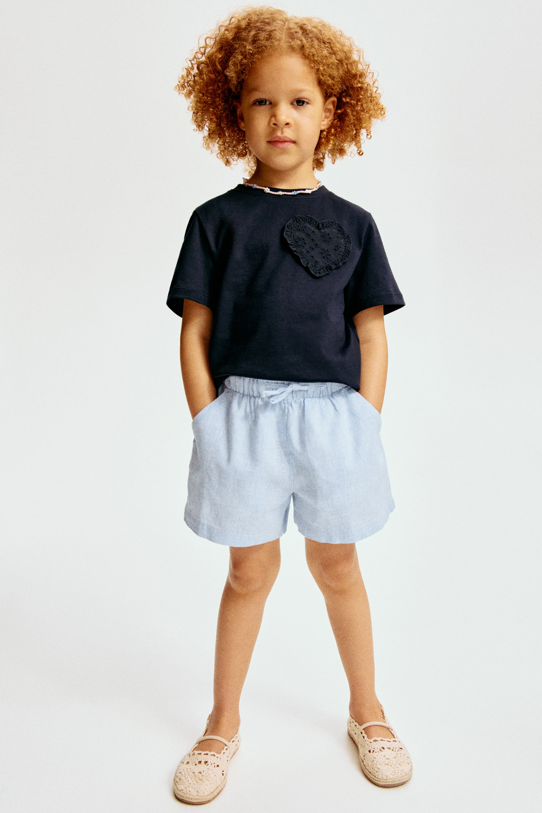 View larger image: This ensemble features a dark navy t-shirt with short sleeves, adorned with a ruffled, beaded neckline trim, and an embroidered heart patch on the left chest. Paired with it are light blue and white striped shorts, designed with an elasticated drawstring waist and convenient pockets. The look is completed with light beige crocheted espadrille shoes, perfect for a stylish and comfortable finish.
