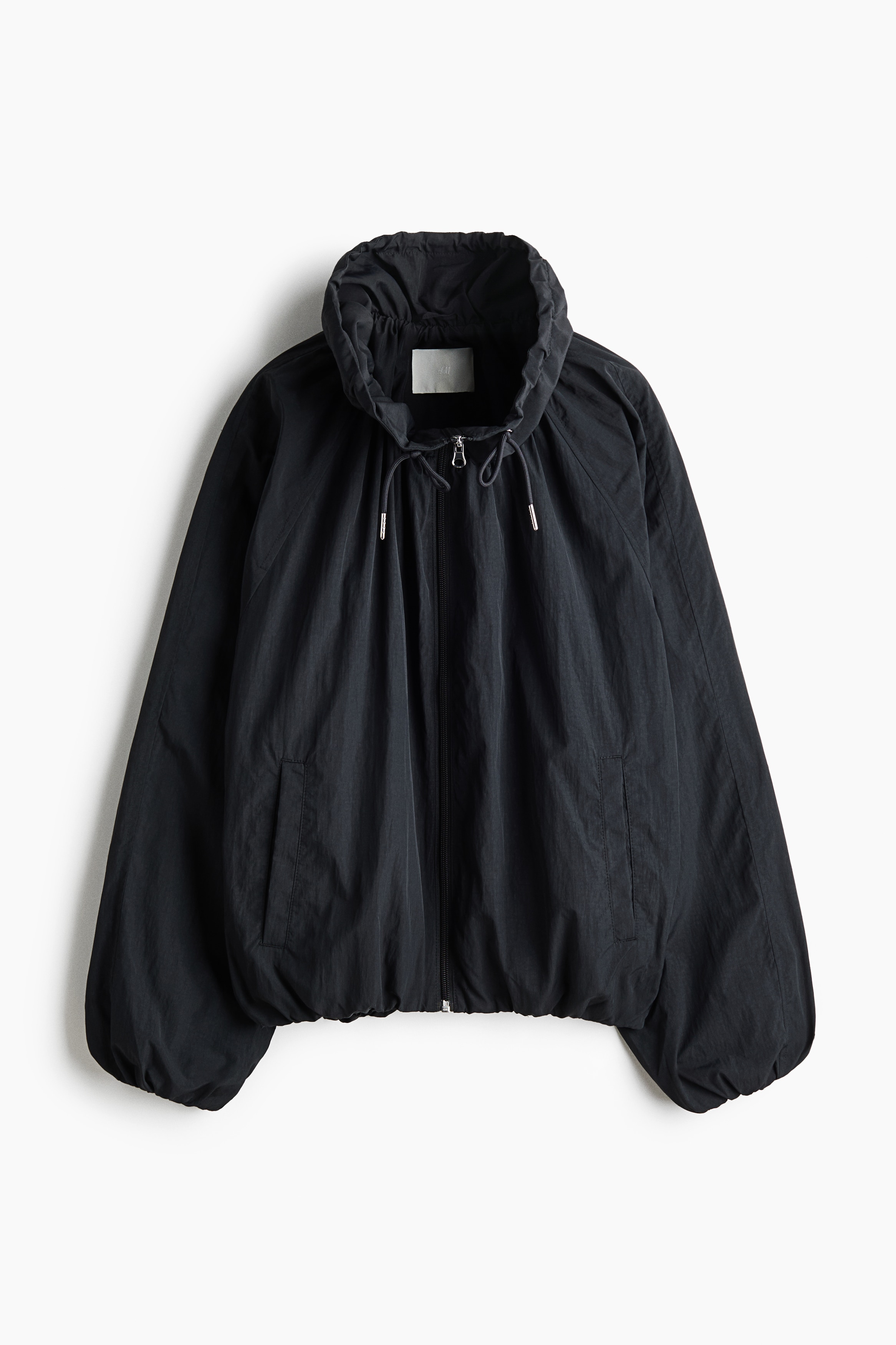 O modelo estÃ¡ a usar uma jaqueta de nylon preta, oversized, com gola alta franzida tipo funil, cordÃ£o ajustÃ¡vel e fecho frontal de correr. A jaqueta tem mangas raglan, criando uma silhueta descontraÃ­da e volumosa.