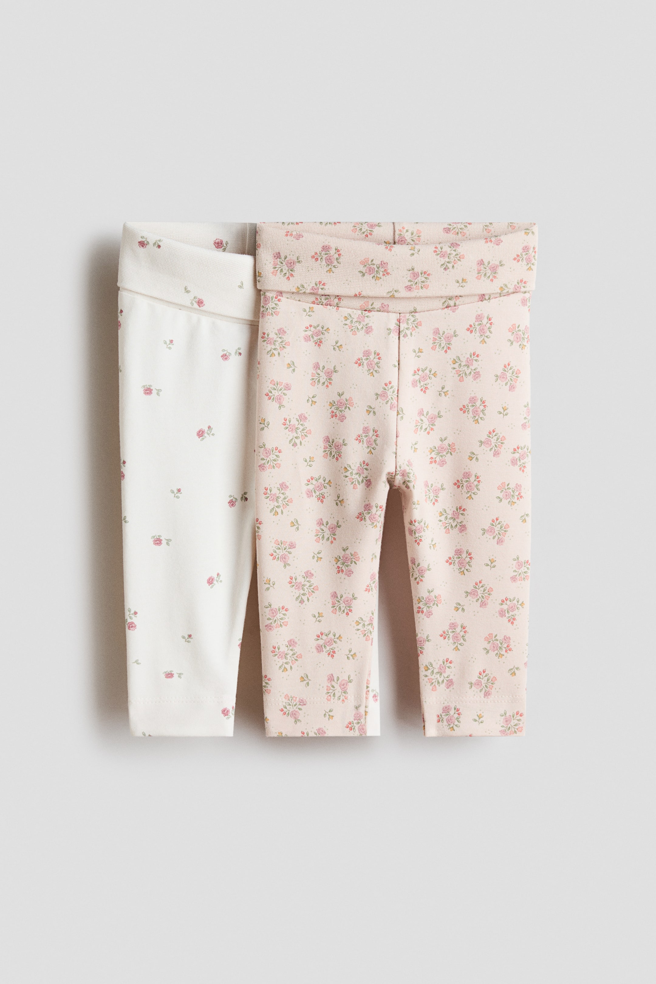 Xem ảnh lớn hơn: Hai chiếc quần legging cho bé được trưng bày từ phía trước. Chiếc bên trái có màu trắng ngà với họa tiết hoa nhỏ màu hồng, trong khi chiếc bên phải có màu hồng nhạt với họa tiết hoa tương tự. Cả hai chiếc quần legging đều có phần cạp gập.