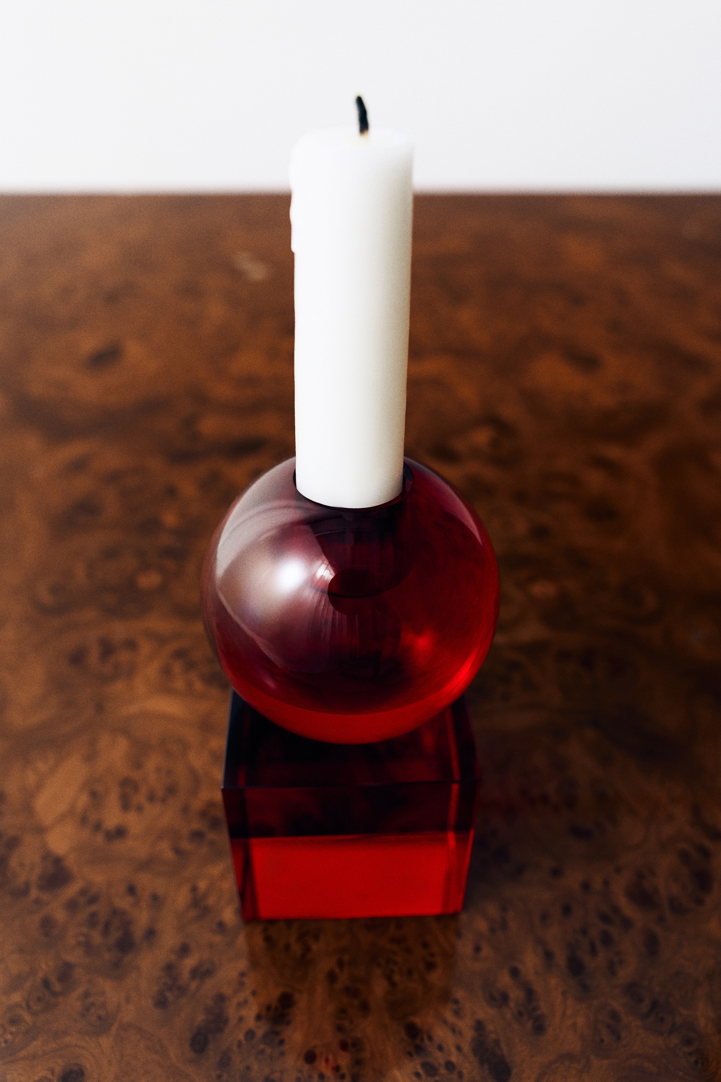Il portacandela in vetro rosso, mostrato frontalmente, presenta una base cubica in vetro rosso traslucido sormontata da una sfera in vetro coordinata. Una candela bianca alta e affusolata Ã¨ posizionata verticalmente, inserita nell'apertura superiore della sfera.