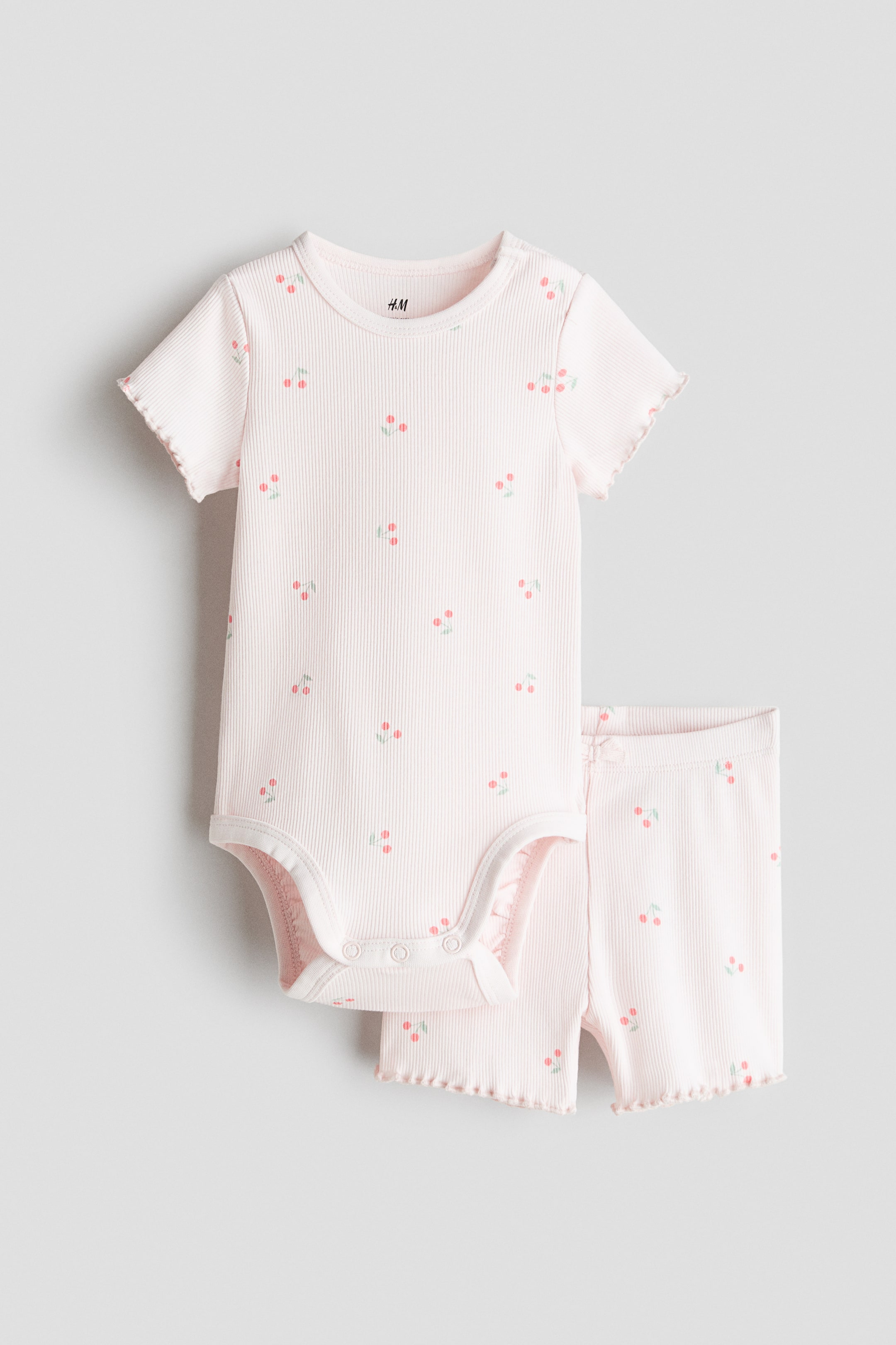 View larger image: A light pink two-piece baby outfit showcases a short-sleeved bodysuit and matching shorts, both crafted from ribbed fabric adorned with a scattered cherry print. The bodysuit features a round neckline, short sleeves with lettuce edge cuffs, and snap closures at the crotch, while the shorts highlight lettuce edge leg openings.