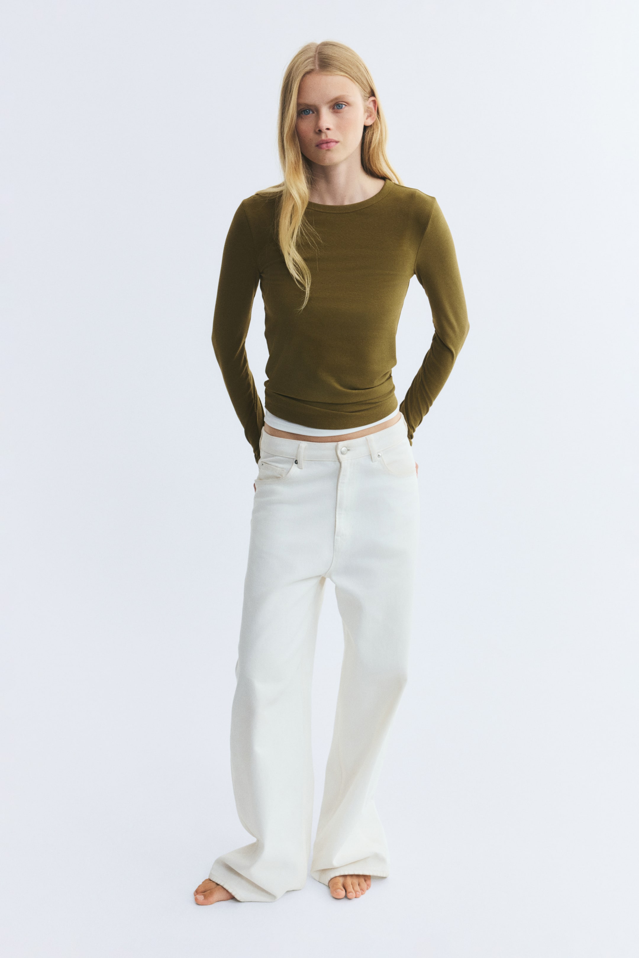 View larger image: The model is wearing a fitted, olive-green long-sleeved top with a round neckline, paired with white wide-leg trousers. The top is styled over a thin white underlayer visible at the hem, as the model stands with hands behind their back.