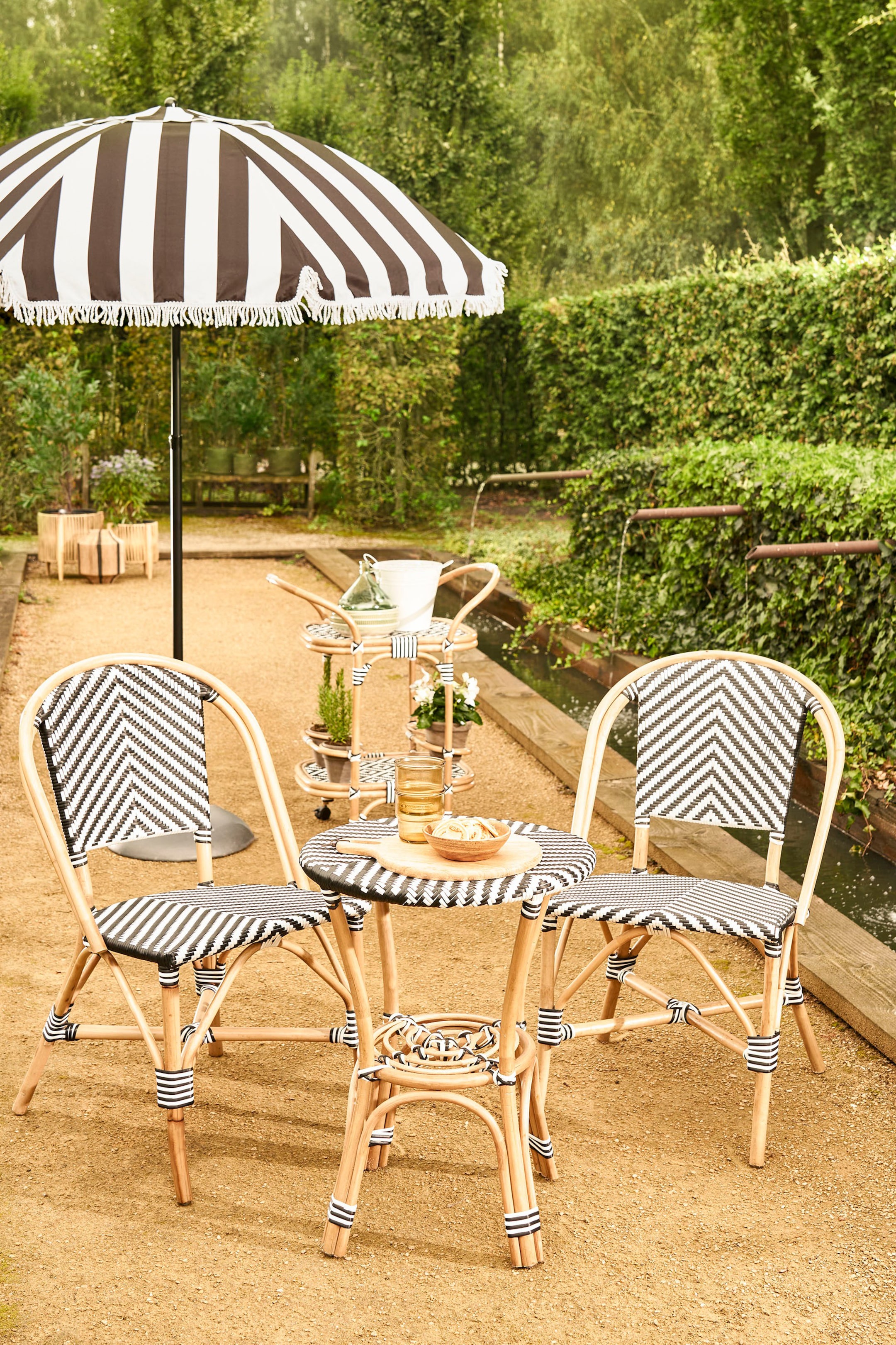 Se større billede: Et udendørs spisesæt med et rundt bord og to stole, fremstillet af lyse rattanrammer. Bordets runde top viser et vævet sort og hvidt stribet mønster, og en lille træskærebræt med snacks hviler på det. Hver stol har et vævet sort og hvidt chevronmønster på ryglænet og et stribet mønster på sædet, med sort og hvidt stribet indpakning, der fremhæver rattanforbindelserne.