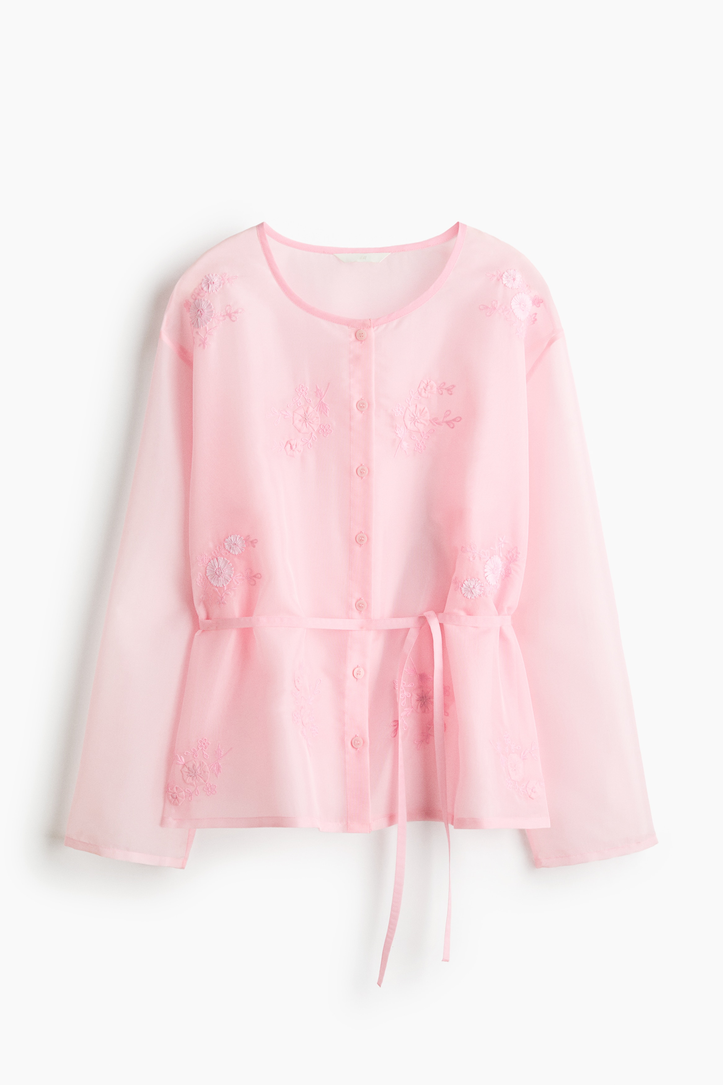 The model is wearing an embroidered organza blouse in light pink, paired with a matching embroidered skirt. The sheer, button-up blouse features delicate floral embroidery on the chest, shoulders and hem, and is styled with a matching slim tie belt at the waist.