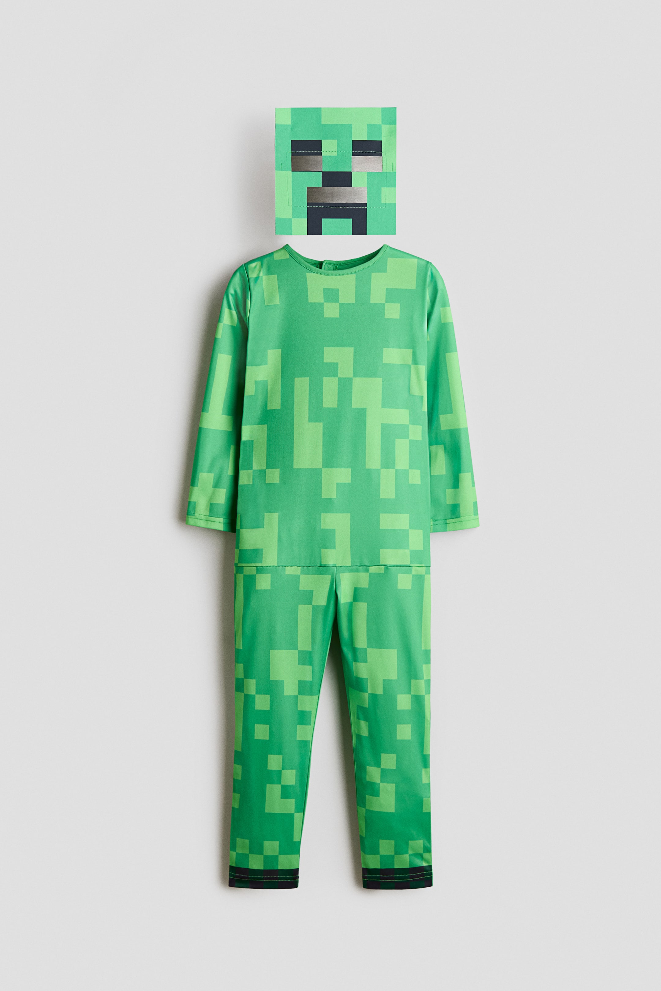Vezi imaginea mai mare: Costum de carnaval: Un costum din două piese verde, pixelat, care include un top cu mâneci lungi și pantaloni lungi asortați, ambele afișând nuanțe variate de pătrate verzi. Costumul este orientat spre înainte, cu o mască pătrată separată, de asemenea în modelul verde pixelat, având decupaje dreptunghiulare întunecate pentru ochi și o deschidere în formă de T pentru gură.