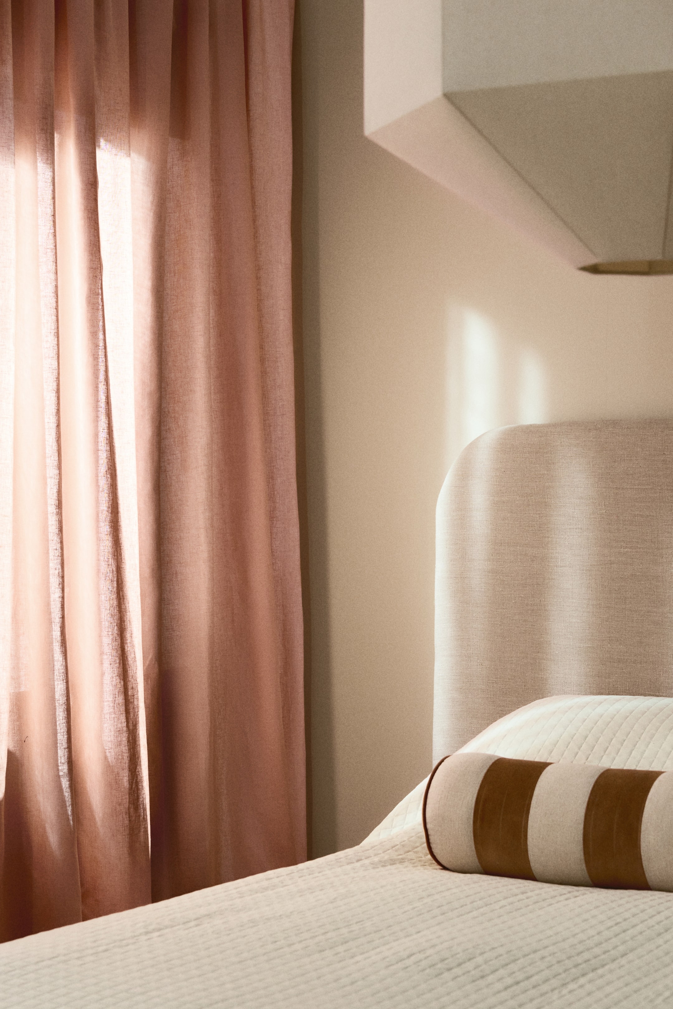 Se større billede: En lysebeige polstret sengegavl med en rundet topkant hæver sig bag et cremefarvet quiltet sengetæppe. En cylindrisk bolsterpude med skiftevis lysebeige og brune striber hviler på sengetæppet, let drejet mod højre.