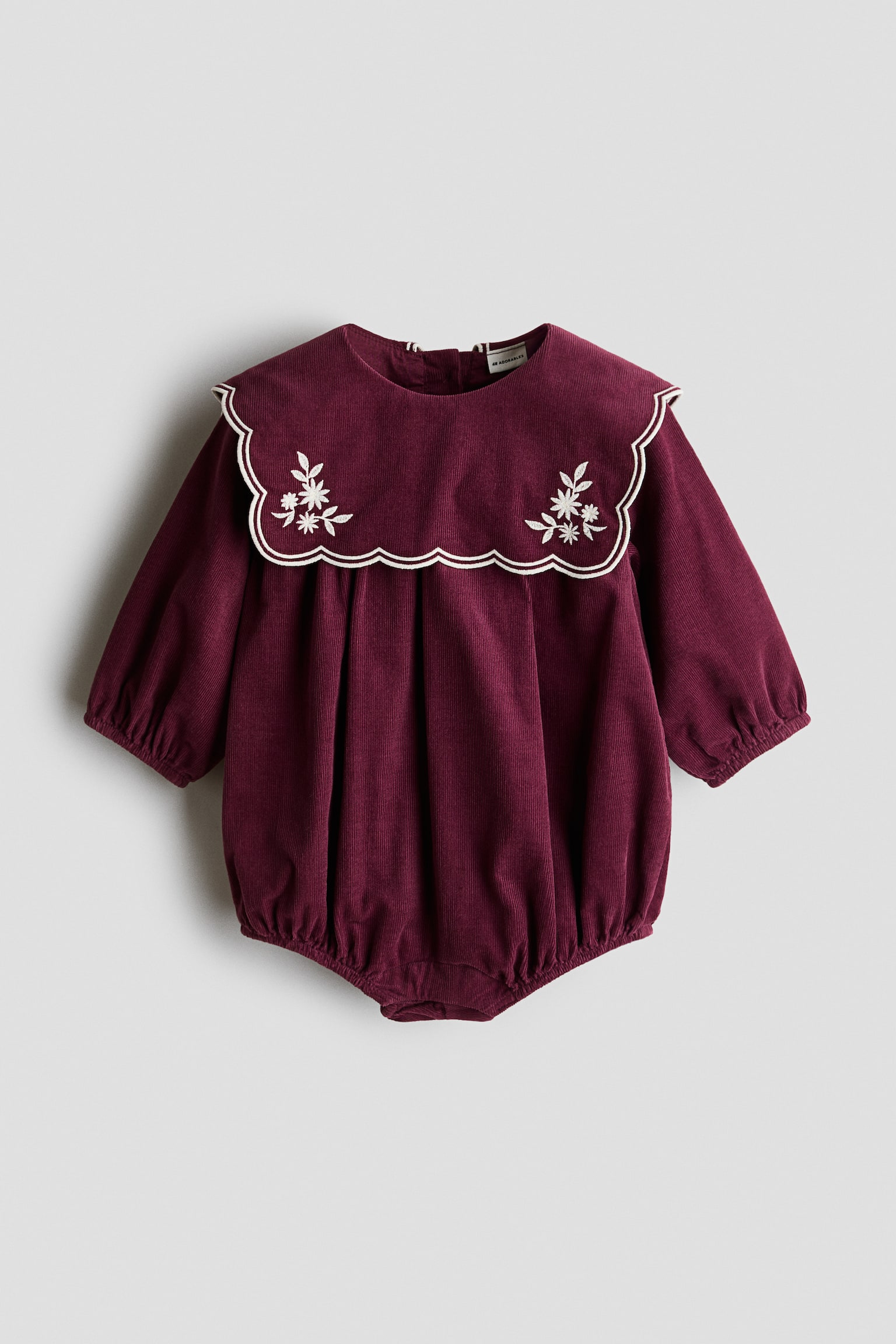 A burgundy corduroy baby romper is shown from the front, featuring a large scalloped collar with white piped trim and white floral embroidery. The romper is designed with long sleeves with elasticated cuffs and a gathered, bubble-style body with elasticated leg openings.