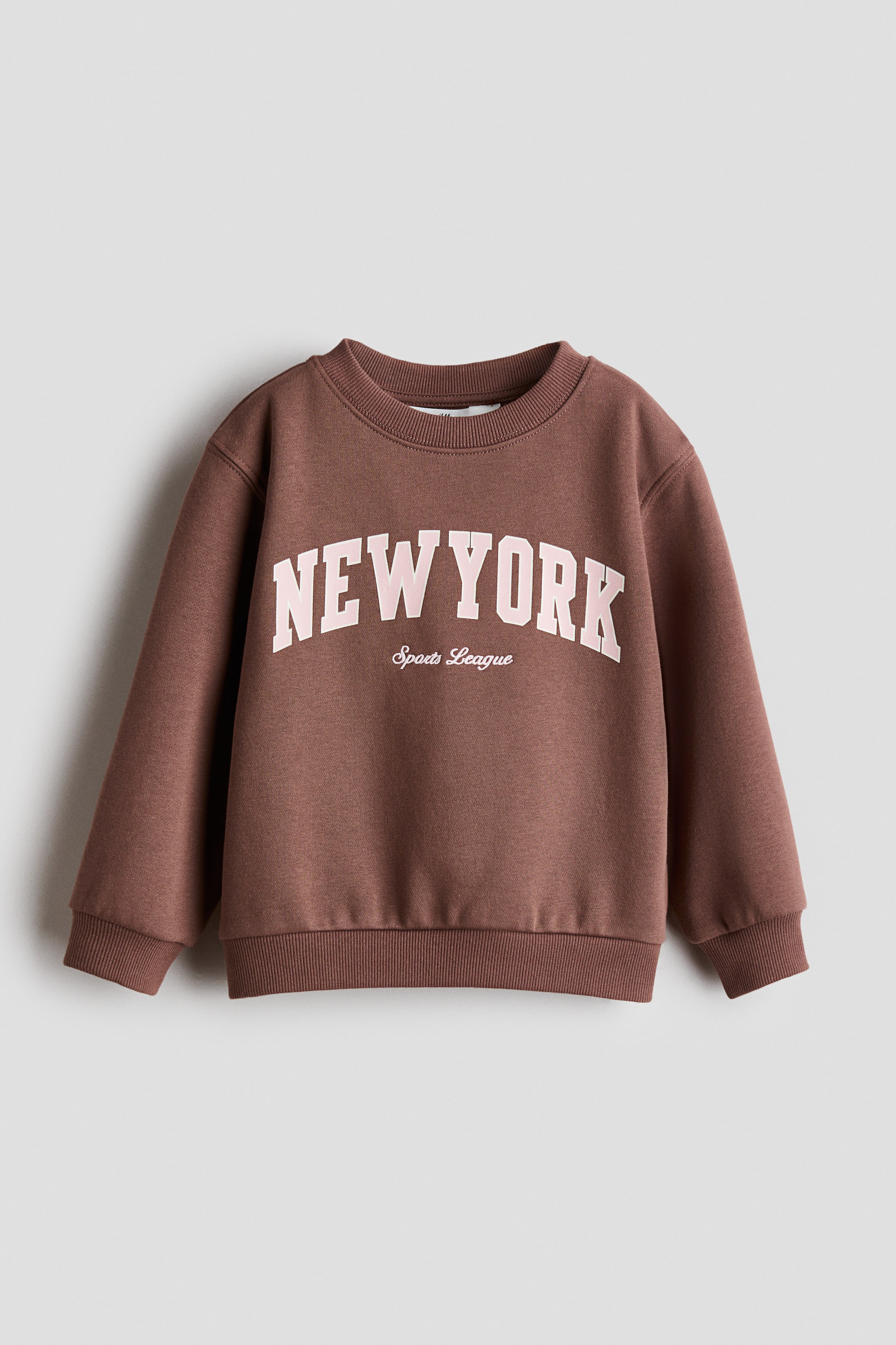 View larger image: A brown crew neck jumper is facing front, featuring "NEW YORK" in arched, light pink block letters across the chest, with "Sports League" in smaller, italicised script below it. It is designed with a ribbed crew neck, ribbed cuffs, and a ribbed hem.