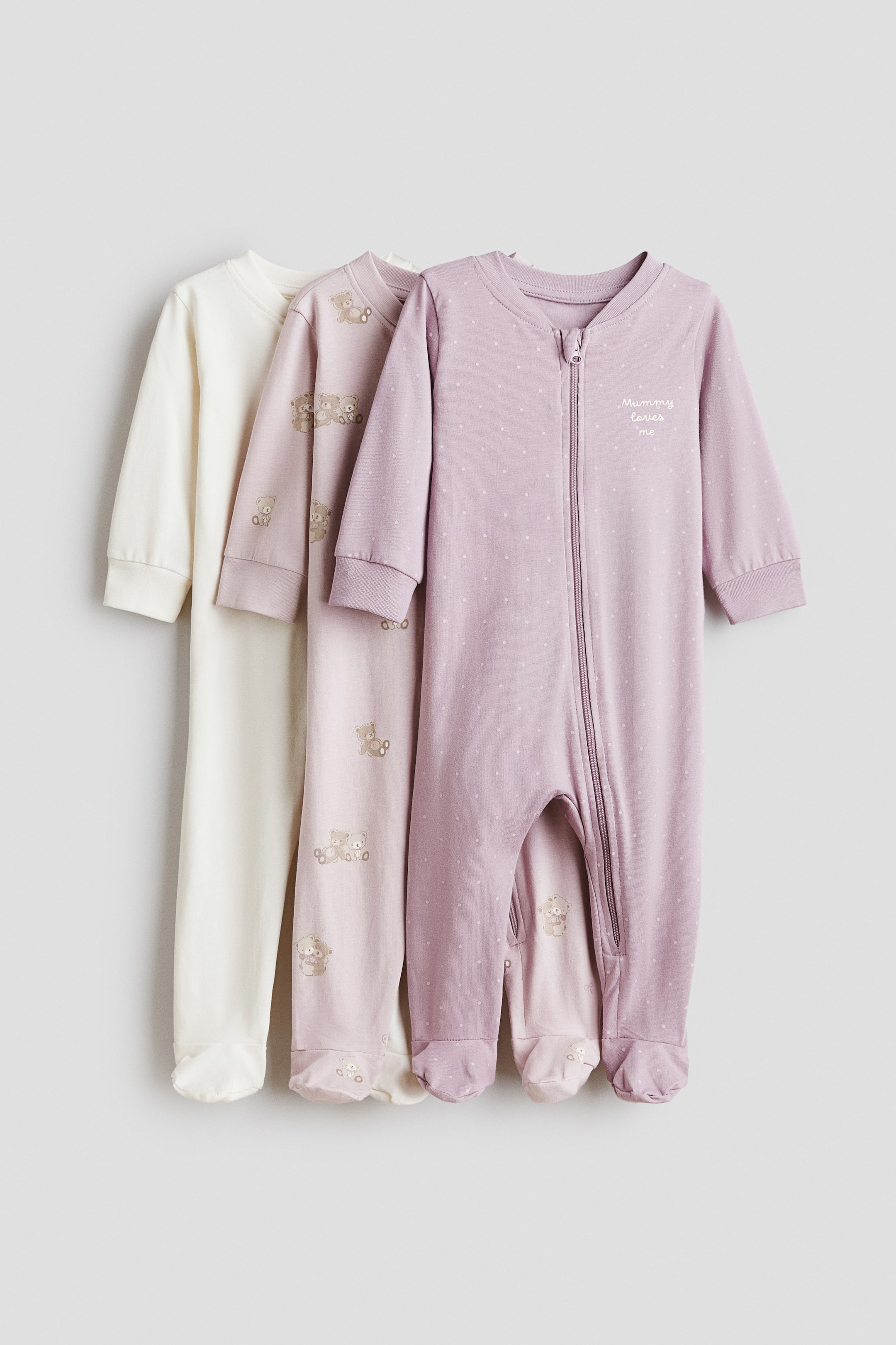 Visa större bild: Tre babypyjamas visas, alla med långa ärmar, ribbade muddar och integrerade fötter. Den vänstra pyjamasen är krämfärgad, den i mitten är ljusrosa med ett övergripande björnmotiv, och den högra är lila med ett allover-prickmönster samt ”Mummy loves me” broderat på bröstet, med en hellång dragkedja.