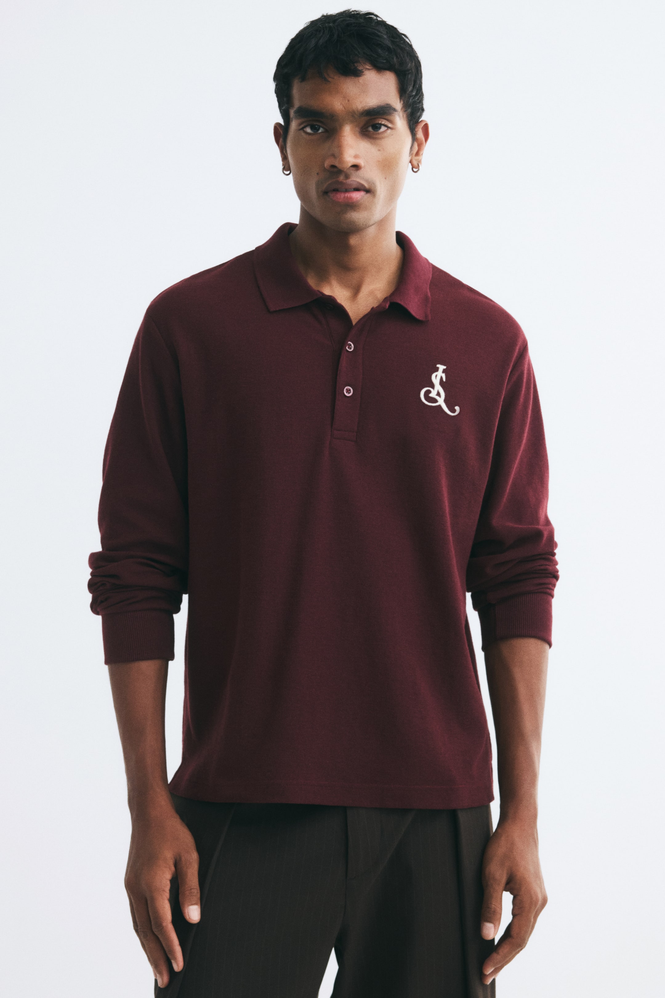 View larger image: The model is wearing a burgundy long-sleeved polo shirt featuring a waffle-knit texture, a ribbed collar, a button placket, and a white embroidered logo on the chest. The shirt has a regular fit and is styled with dark trousers.
