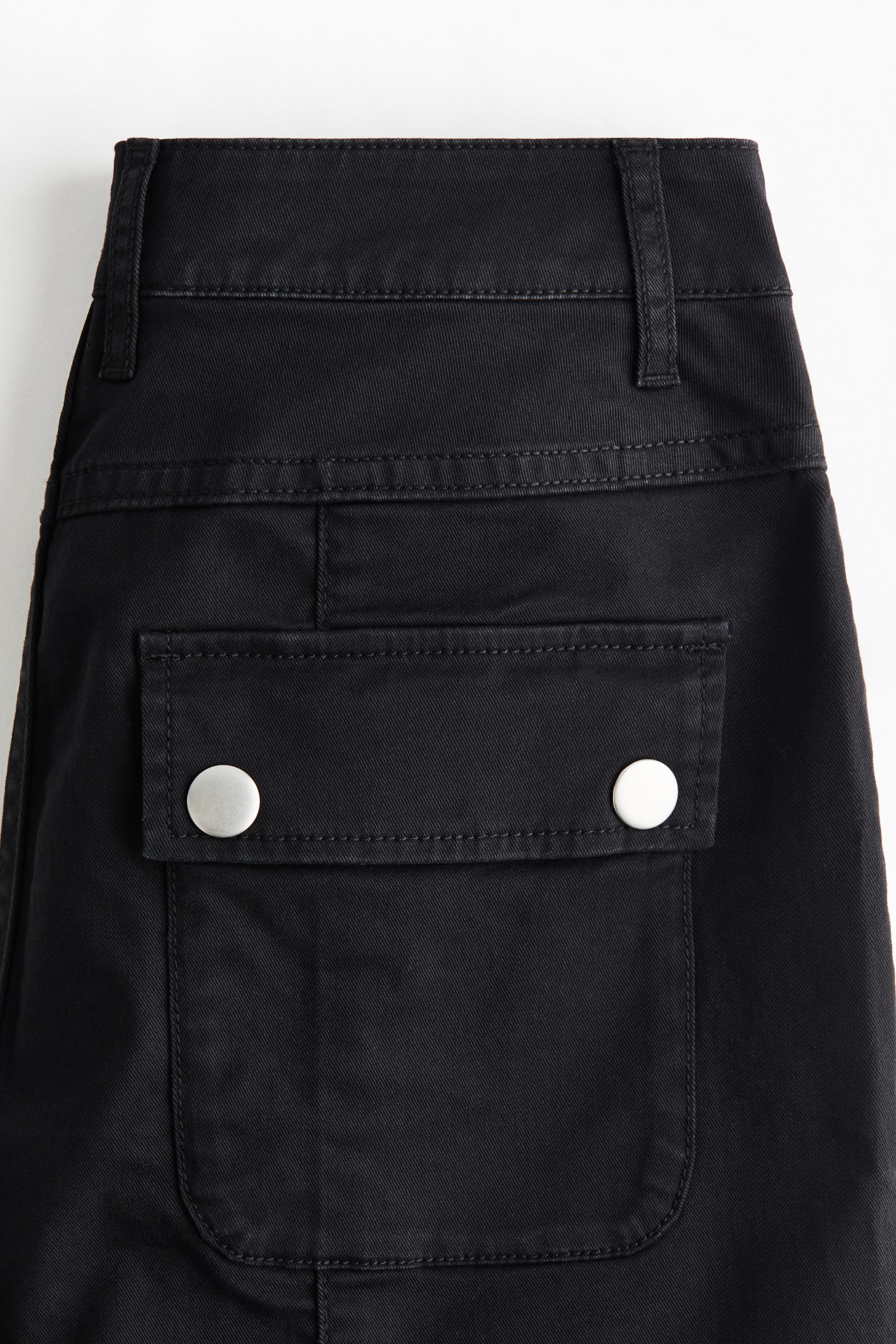 Xem ảnh lớn hơn: Quần đen có đặc điểm nhìn từ phía sau với cạp quần rộng, nhiều đai giữ dây lưng, và một túi nắp hình chữ nhật ở giữa được cố định bằng hai nút nhấn màu bạc.