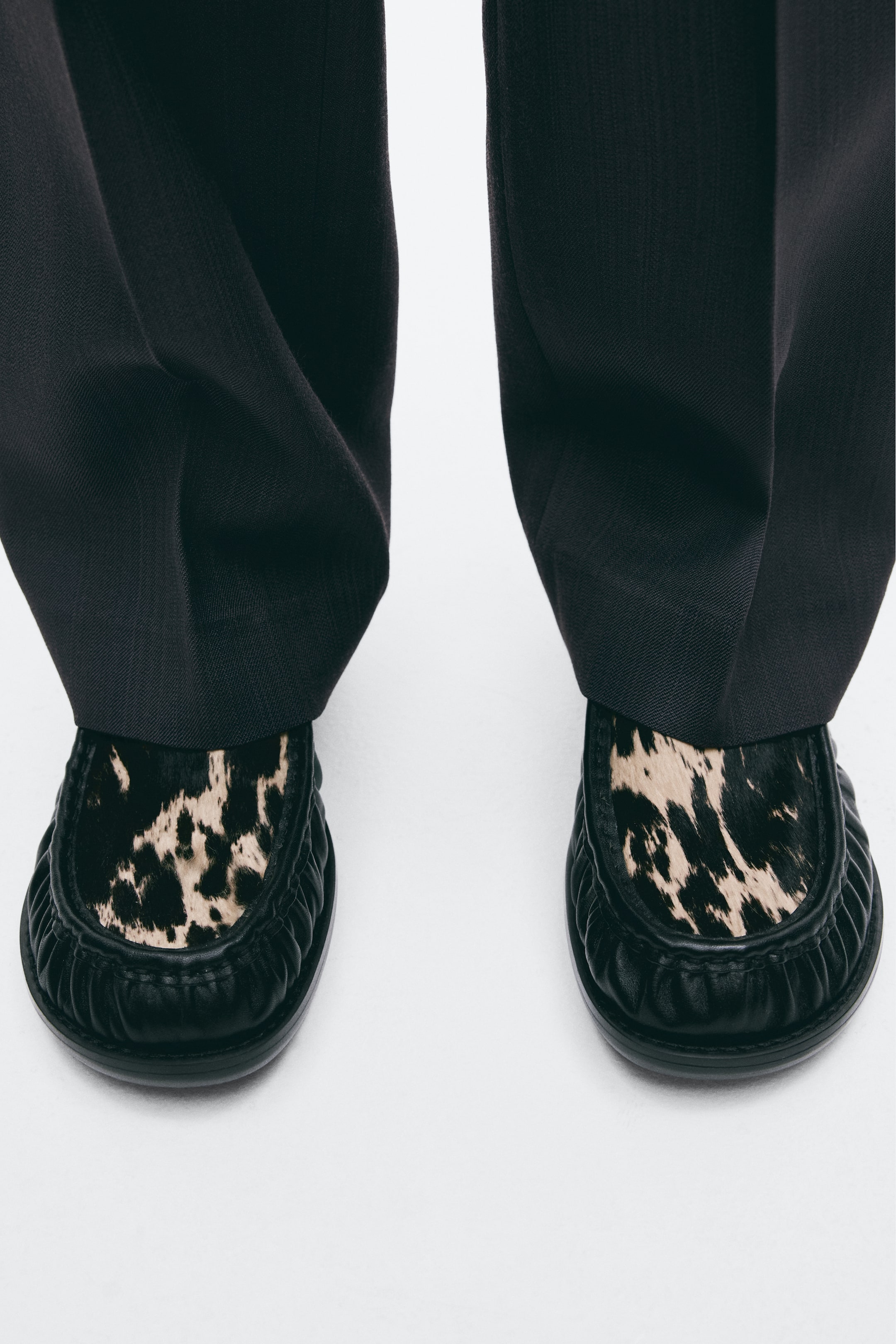 Visa större bild: Svarta läderskor av loafertyp med en rynkad mokasinsöm och en texturerad ovandel med djurmönster bärs av modellen. Skorna stylas med mörka byxor med vida ben som faller lätt över skornas ovandelar.