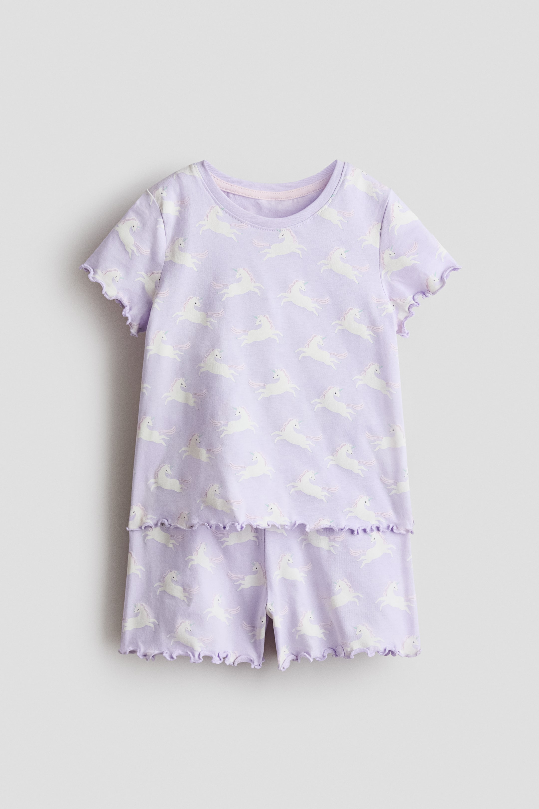 View larger image: A pair of light purple pyjamas is shown from the front, crafted from cotton jersey and featuring a repeating white unicorn pattern. The short-sleeved top presents a round, light-pink rib-trimmed neckline and overlocked, ruffled edges at the short sleeves and hem, while the matching shorts feature an elasticated waist and overlocked, ruffled hems.