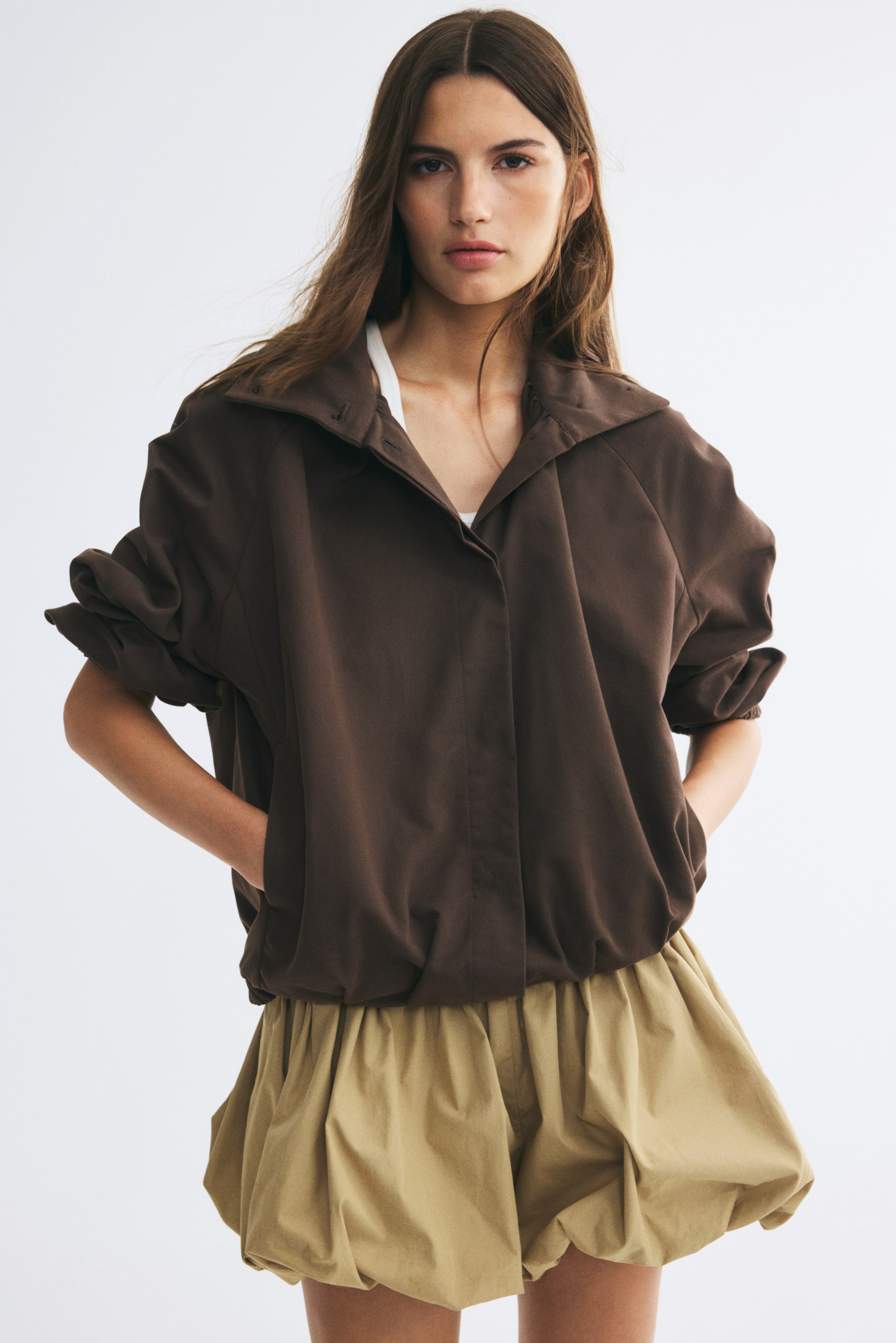 Zobrazit větší obrázek: Modelka má na sobě tmavě hnědou oversized keprovou bundu a stojí s rukama zasunutýma do bočních kapes. Bunda má otevřený stojáček, vyhrnuté raglánové balónové rukávy a elastický balónový lem, je stylizovaná přes bílé top a doplněná béžovými balónovými šortkami.