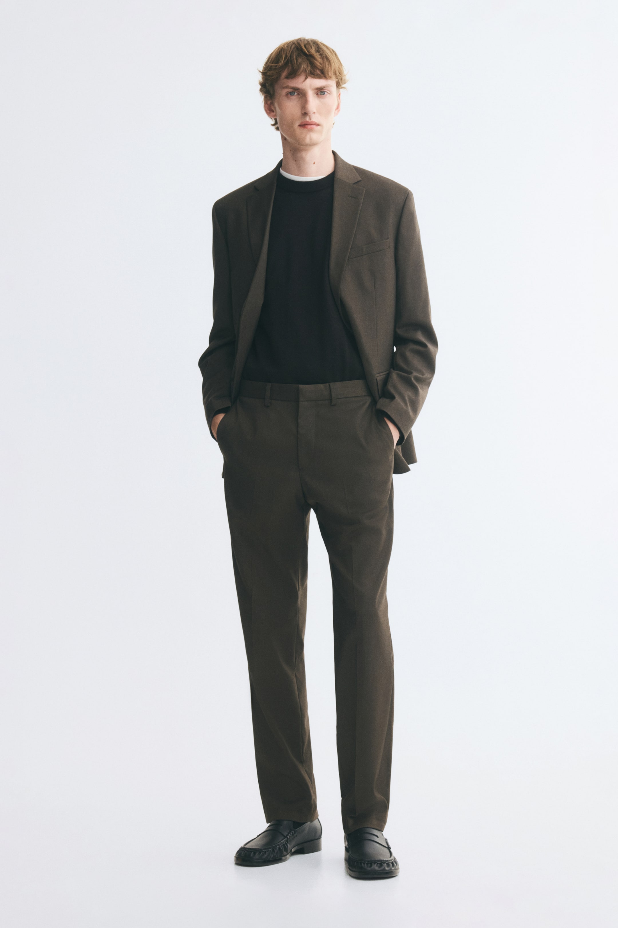 View larger image: A dark brown suit, comprising a single-breasted blazer and matching tailored trousers, is worn by the model with hands placed in the trouser pockets. The suit is layered over a black crewneck top and paired with black leather loafers.