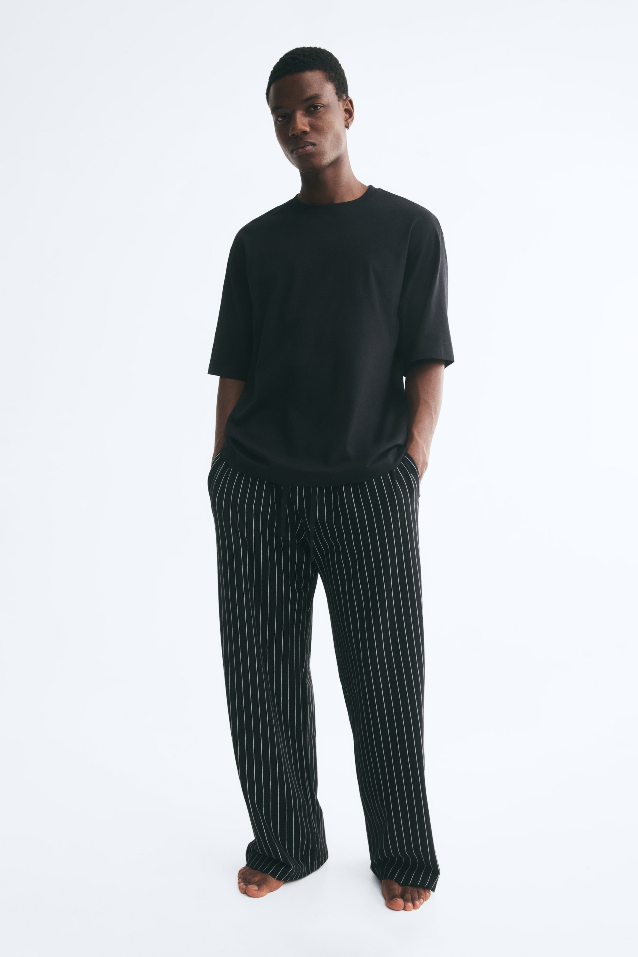 Agrandir l'image: Un ensemble de pyjama composé d’un t-shirt noir uni à coupe décontractée, avec un col rond, et d’un pantalon noir assorti à coupe droite, orné de fines rayures blanches verticales, est porté par le mannequin. Le mannequin est debout, pieds nus, les mains dans les poches latérales du pantalon.