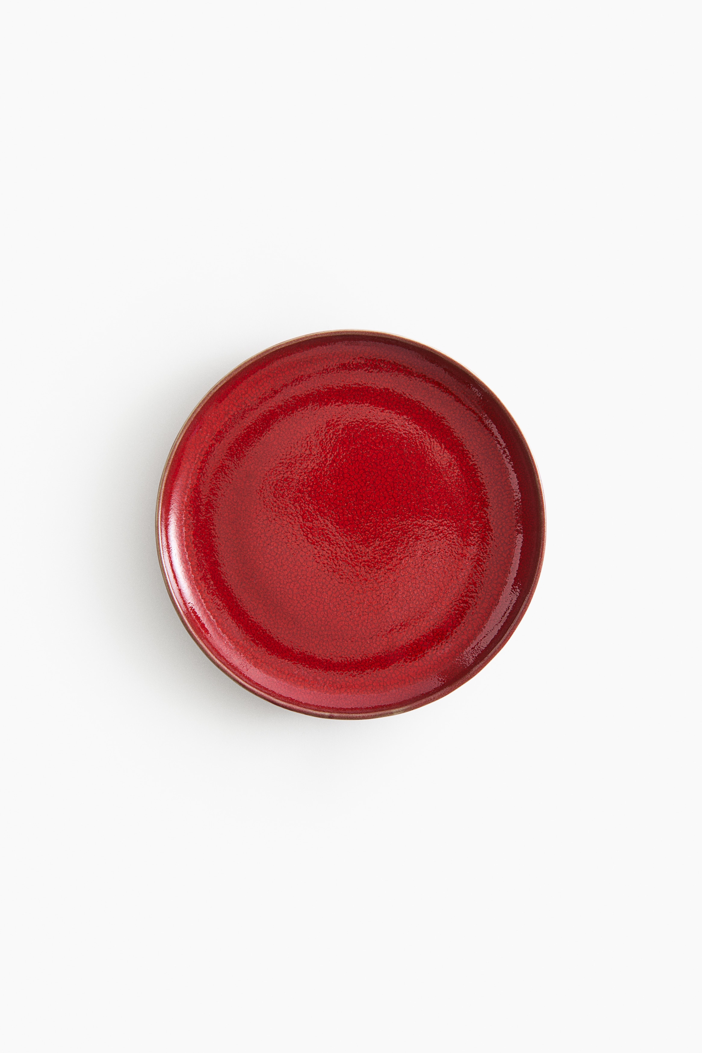 Une assiette ronde en céramique rouge brillante, dotée d’un émail craquelé, tient une fourchette en argent poli posée en diagonale sur sa surface. La fourchette présente quatre dents orientées vers le coin supérieur gauche, avec son manche s’étendant vers le coin inférieur droit.