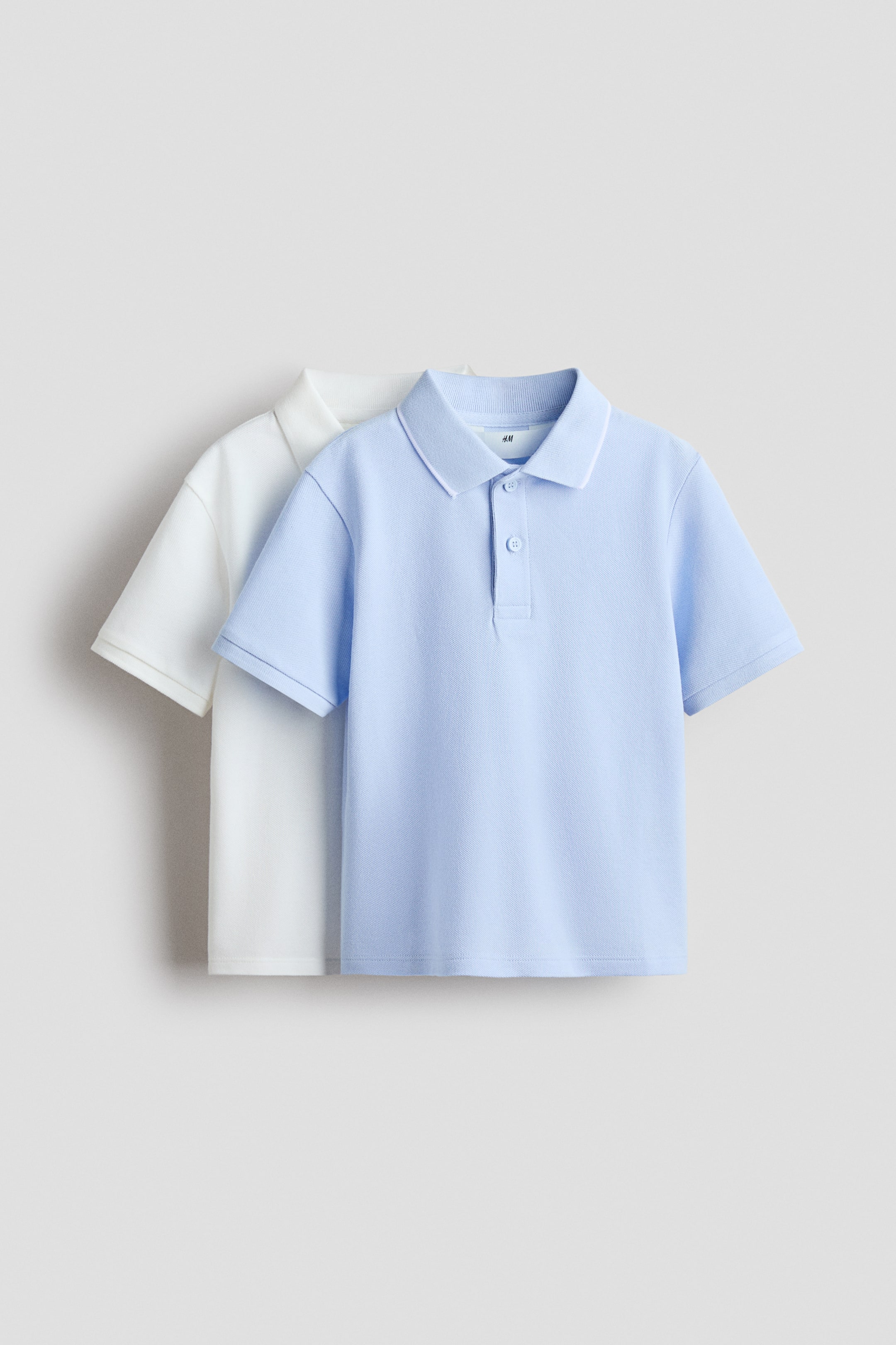 View larger image: Two polo shirts are displayed: a light-blue shirt in the foreground and a white shirt layered behind. Both shirts feature a ribbed polo collar, a two-button placket, and short sleeves with ribbed cuffs.