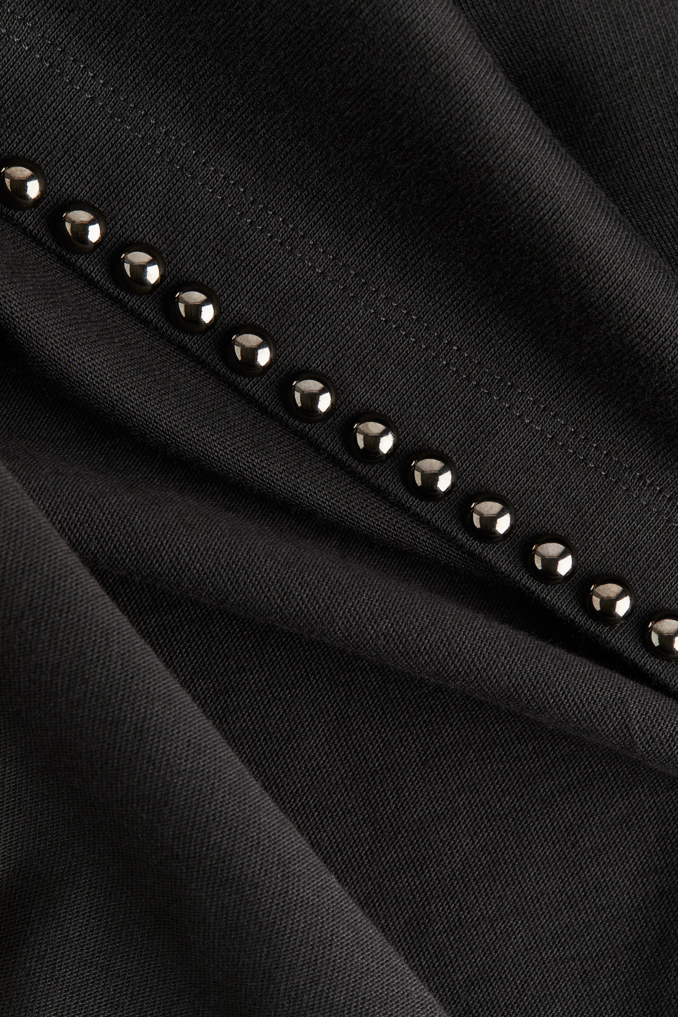 View larger image: A black knit fabric is draped, forming soft folds. A diagonal line of round, metallic studs is running across the fabric, parallel to a visible stitched seam.