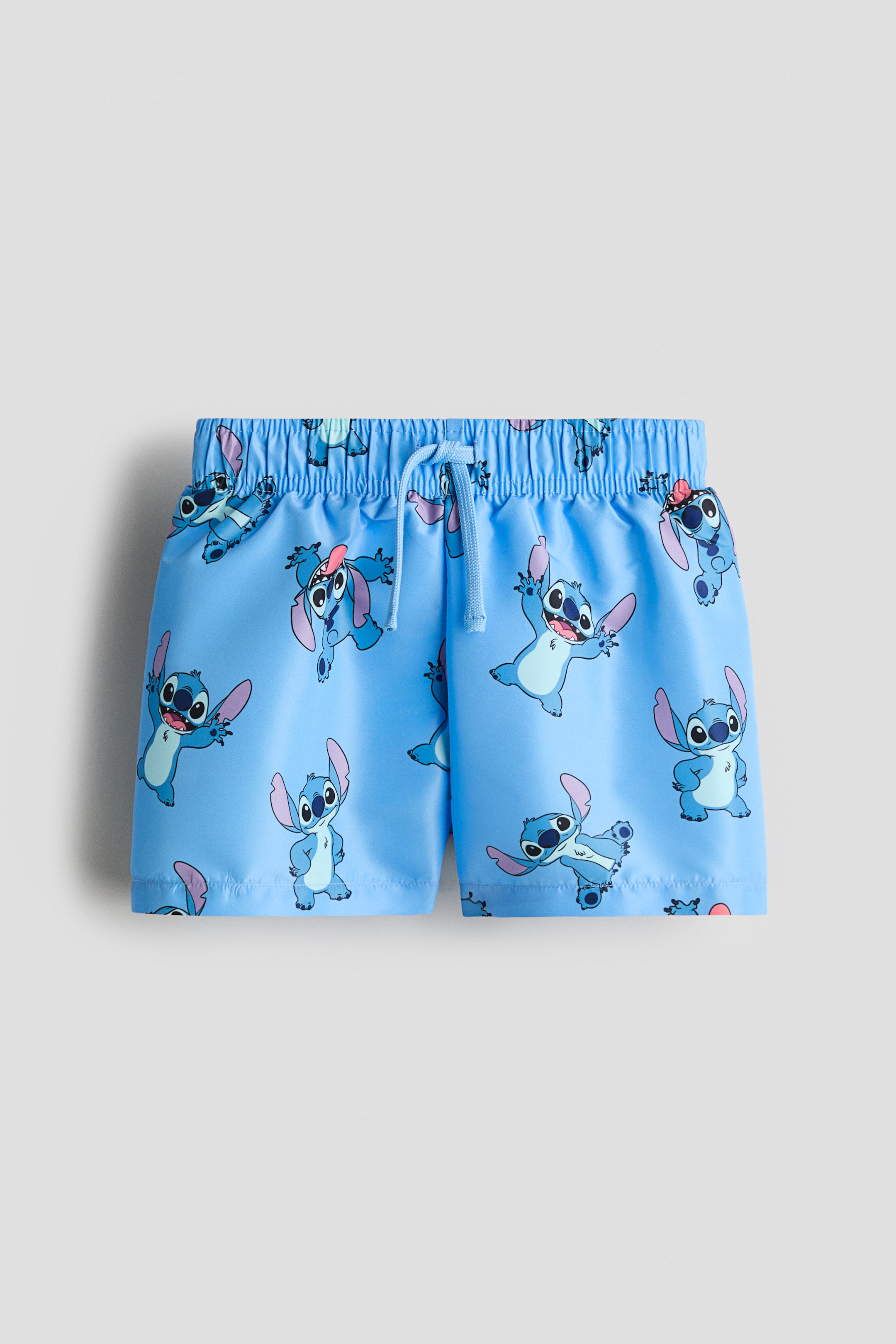 Visualizza immagine più grande: I pantaloncini da bagno frontali presentano un colore azzurro chiaro con un motivo a stampa ripetuto di Stitch. Sono dotati di una vita elasticizzata coperta con un laccetto azzurro chiaro.