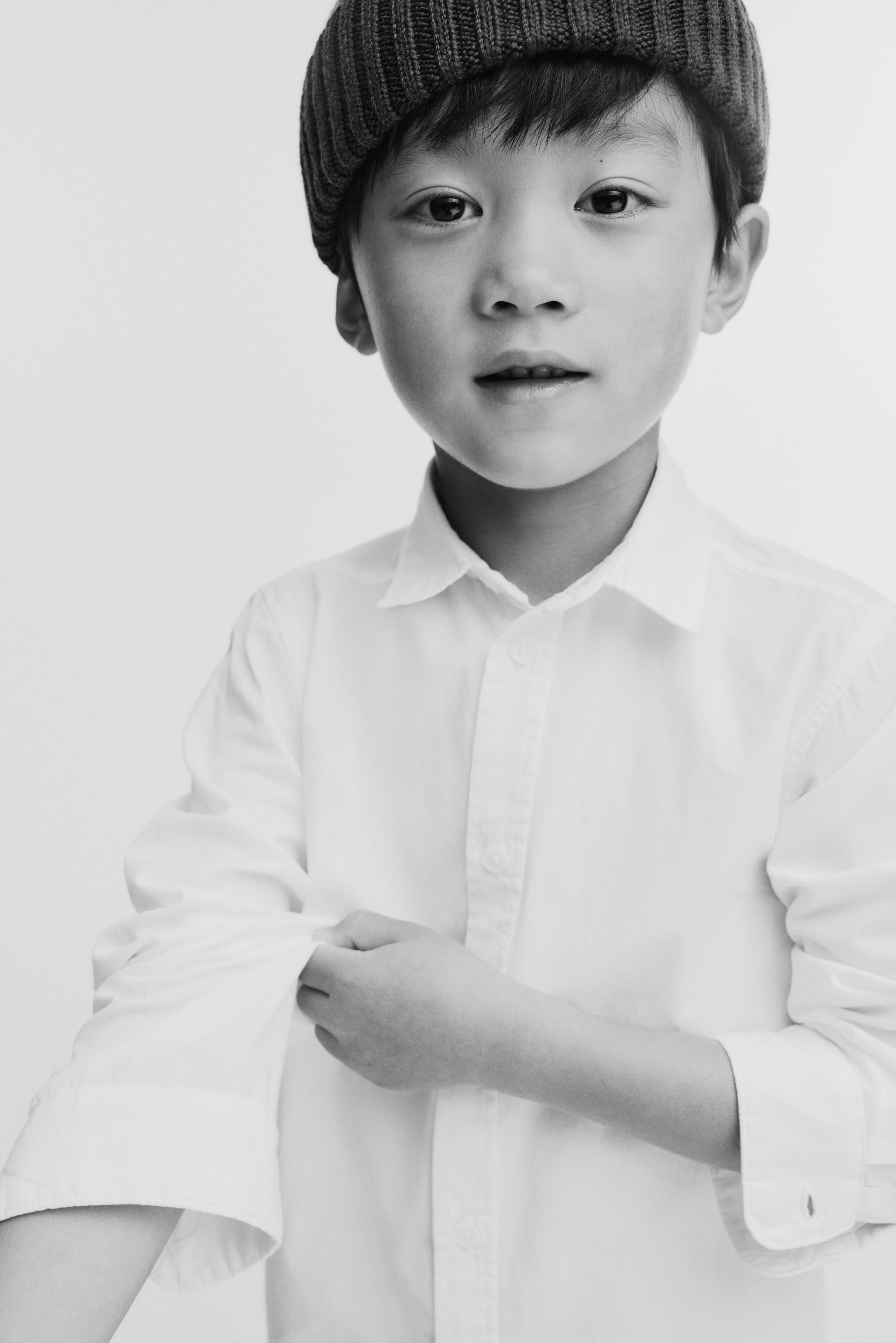 View larger image: A dark, ribbed knitted beanie is fitting snugly on the head, while a white button-up shirt is displaying a pointed collar and a buttoned front placket, with one long sleeve being rolled up at the cuff.