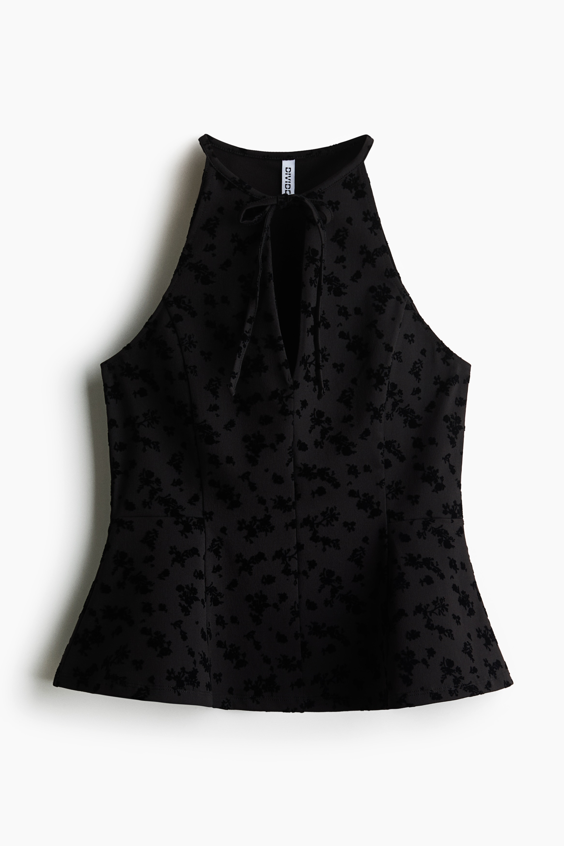 La modelo lleva un top entallado y sin mangas con estampado flock en toda la prenda, que presenta un cuello alto y estrecho con una profunda abertura delantera en forma de V y delicados lazos. El top cuenta con costuras de corte estructuradas y un bajo peplum acampanado, mientras la modelo sostiene las manos cerca del cuello.