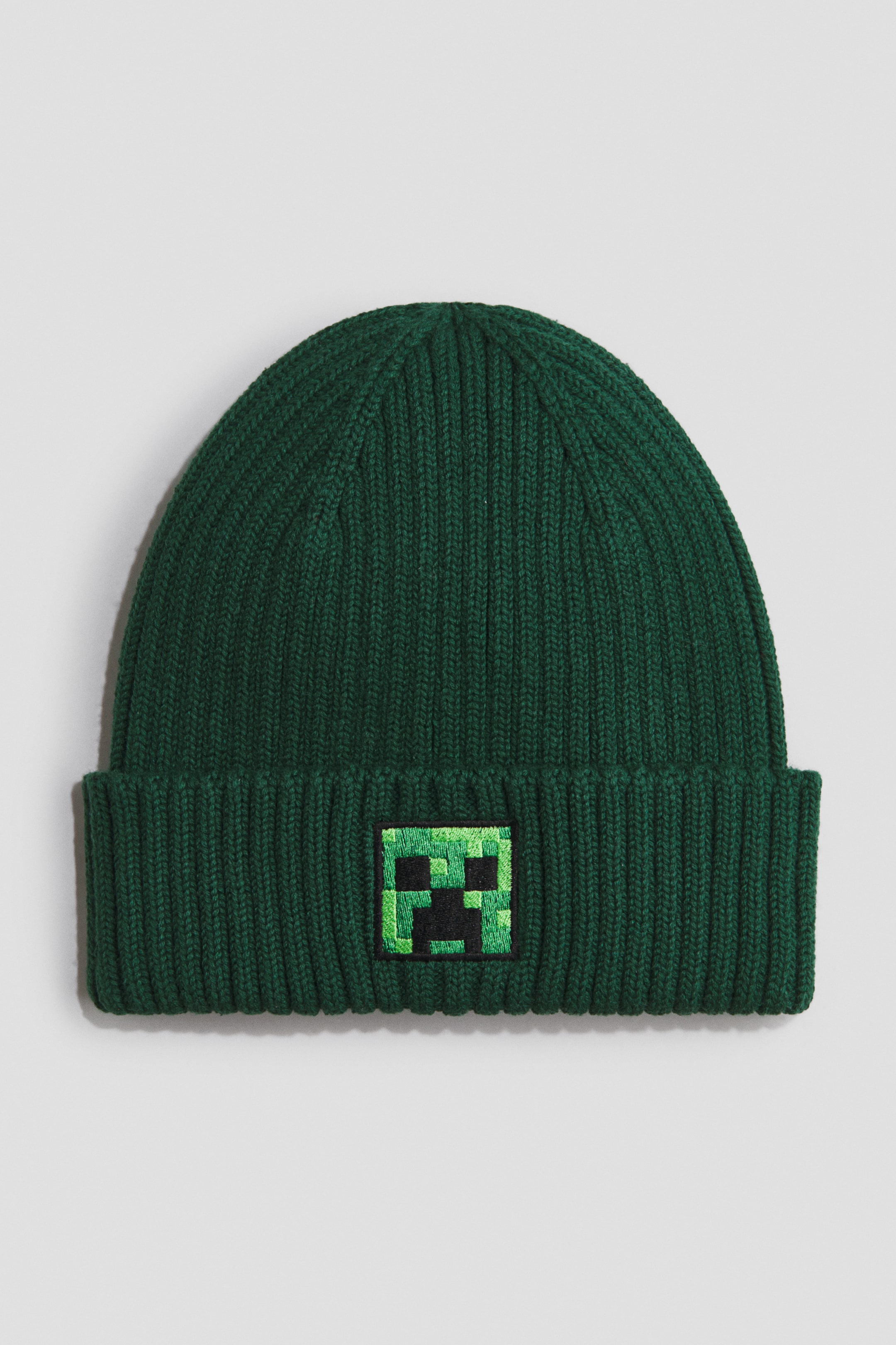 View larger image: A dark green ribbed knit beanie is shown facing forward, featuring a folded cuff. An embroidered square patch, depicting a pixelated green and black face, is centred on the front of the cuff.