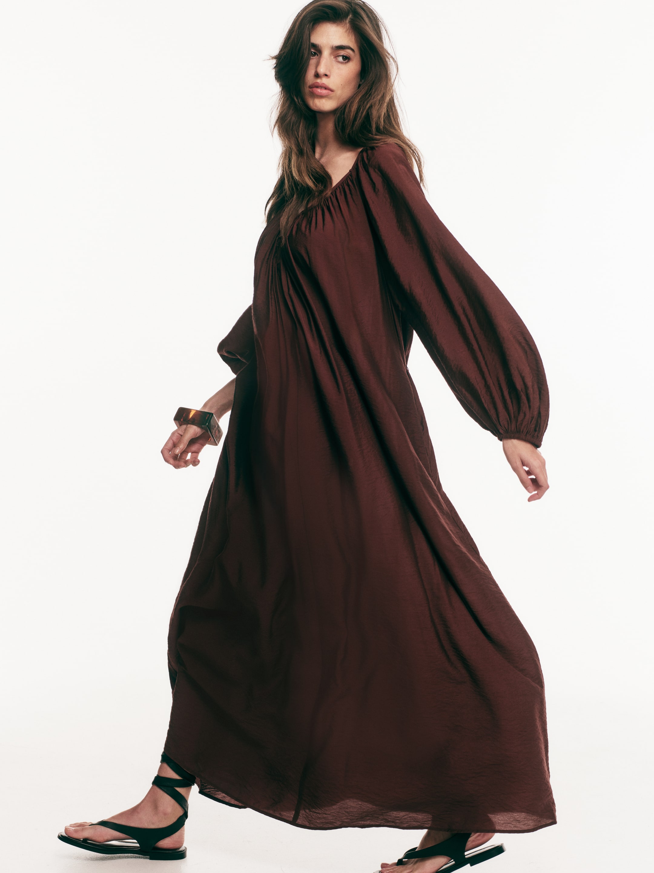 View larger image: The model is wearing a long, oversized tunic dress crafted from a dark reddish-brown woven fabric. It boasts a gathered sweetheart neckline and raglan balloon sleeves with elastic cuffs, while its full skirt flows gracefully around her as she moves.