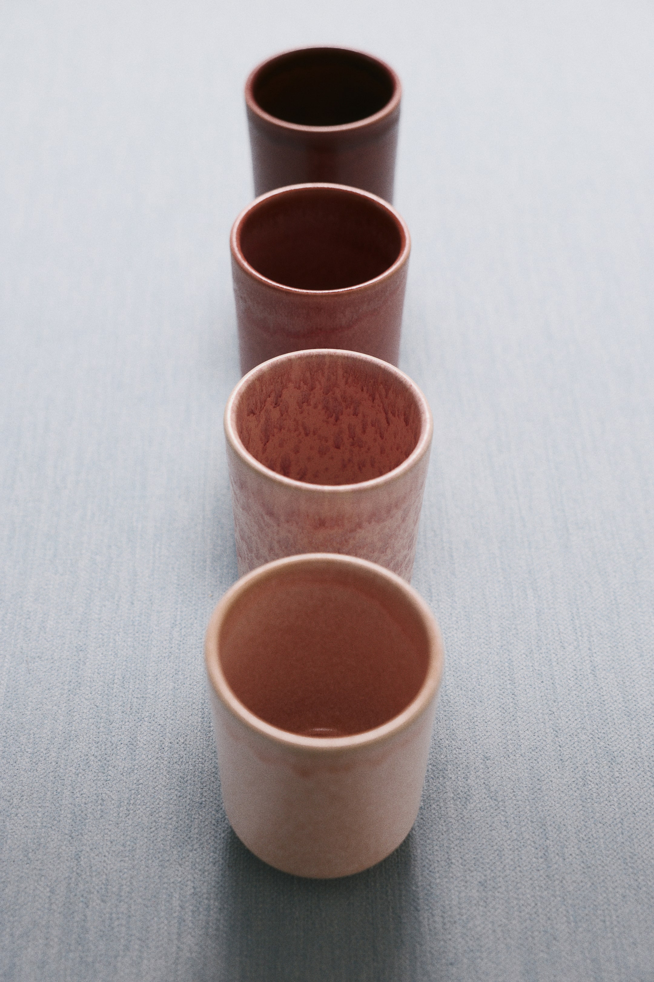 View larger image: Four handleless stoneware cups are standing in a vertical line, showcasing a gradient of glazes ranging from light peach to dark brown. The cylindrical cups are displaying unique textured finishes, including smooth satin surfaces and mottled, speckled patterns on their exteriors and interiors.