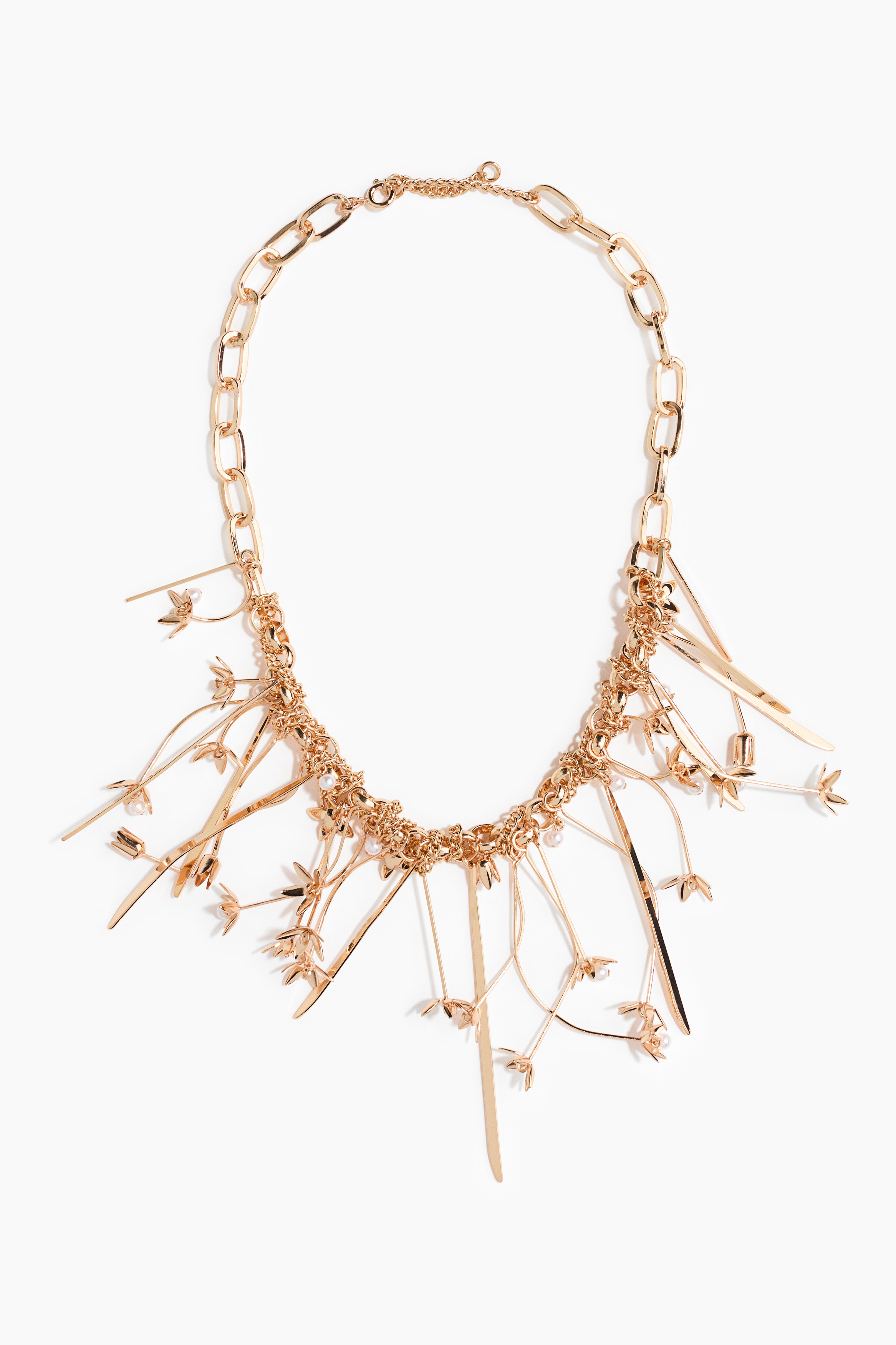 Een opvallende metalen ketting in goudtint, met een schakelketting en een cluster van sierlijk afhangende bloemvormige hangers met kleine parelachtige kralen, wordt gedragen door het model. De ketting is gestyled over een crÃ¨mekleurige trui met hoge hals.